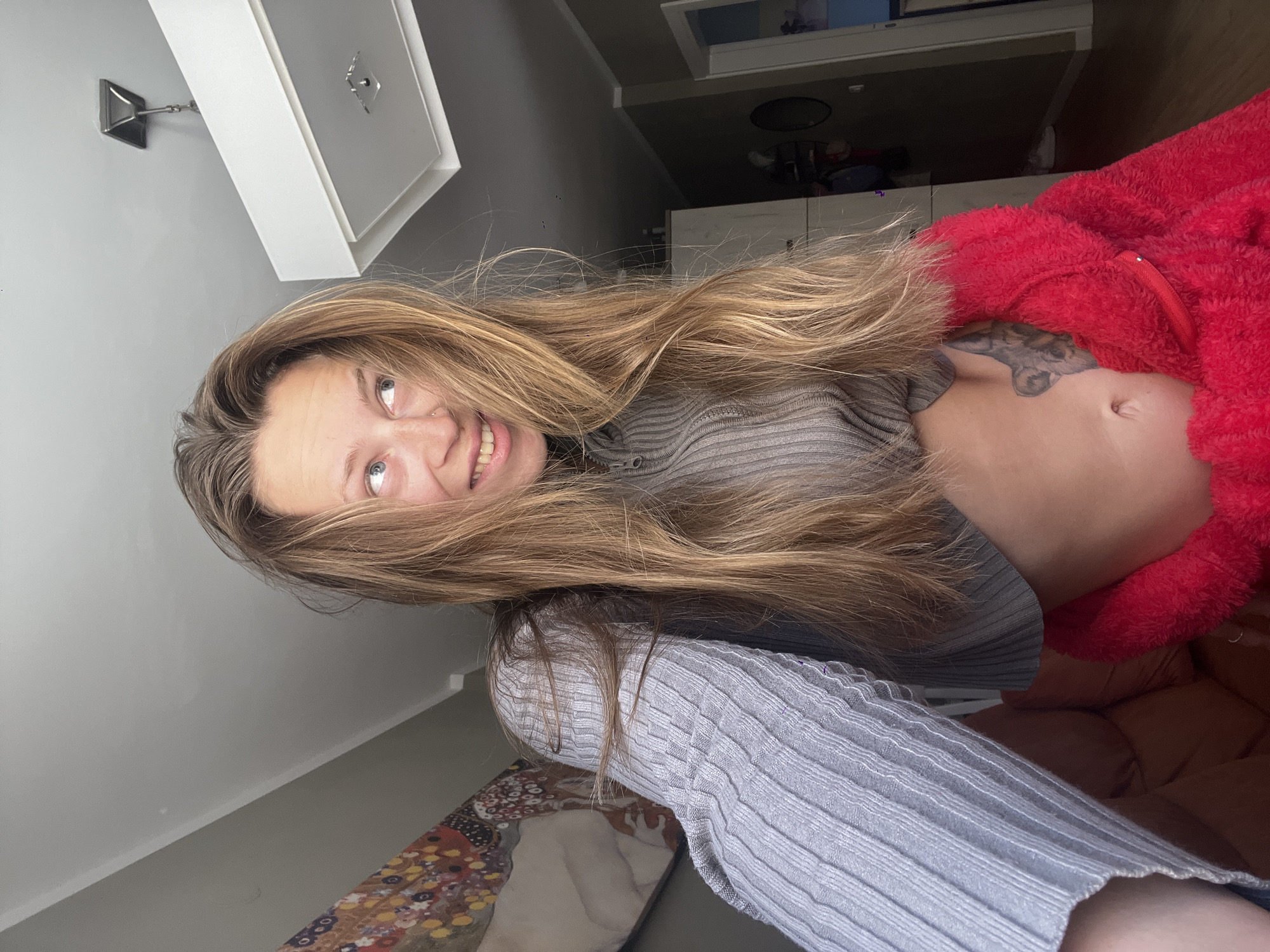 A woman with long, blonde hair is smiling into the camera. She is wearing a grey ribbed sweater and a red fuzzy jacket. Her midriff is exposed, revealing a tattoo on her torso. The background includes a grey wall, a white cabinet, and a partial view of an abstract patterned object at the bottom of the image. A ceiling light fixture is visible on the top left of the frame. The location in the city of Bečići, Montenegro cannot be determined from the image.