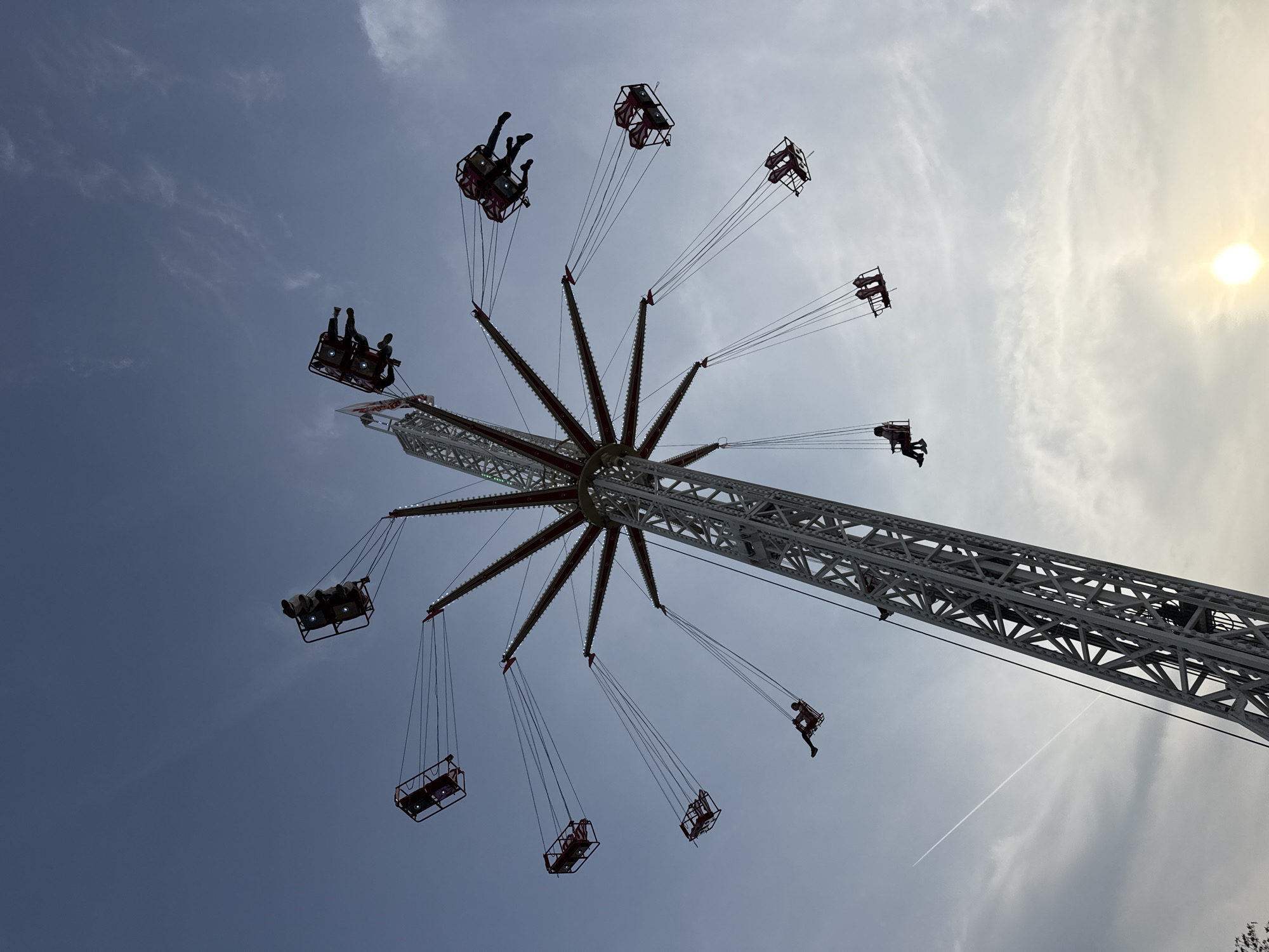 An upward-angle photograph shows a tall, amusement park swing ride in Plovdiv, Bulgaria, set against a blue sky with some clouds and the sun shining brightly. The ride has a central, white, lattice-work tower extending diagonally from the lower right toward the upper left. Around the top of the tower, the ride features a red-and-white star-shaped structure with swings suspended by cables from each point. Most of the swings are occupied by people, some appearing as pairs. A faint contrail from an airplane is visible in the sky near the base of the tower.