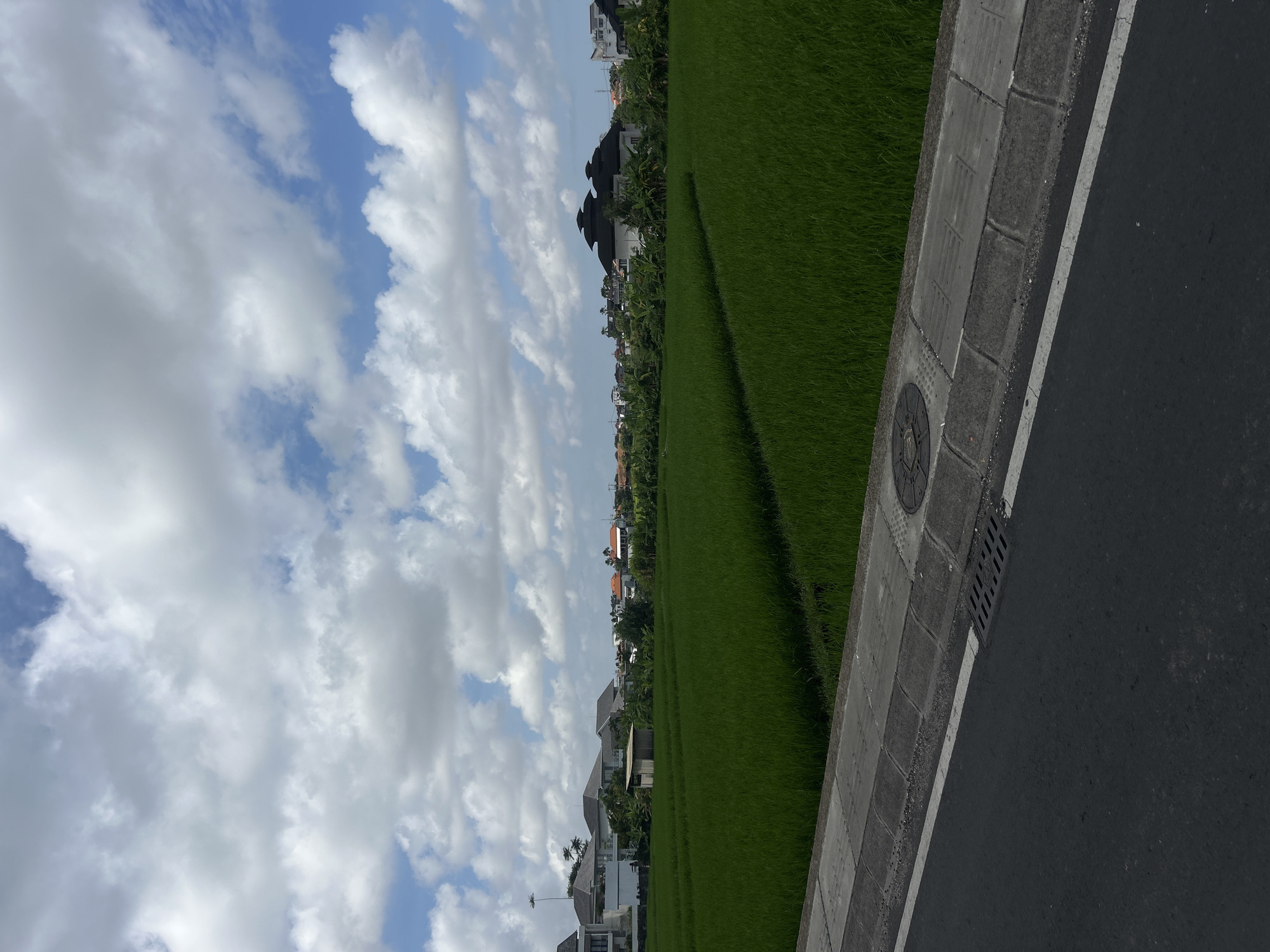 A low-angle, full outdoor shot captures a lush green rice paddy field adjacent to a paved road in North Kuta, Indonesia. The field fills the middle ground and extends to the horizon, where it meets a line of buildings and trees. In the foreground, the asphalt road is visible with a white line marking its edge. A concrete sidewalk with a drainage grate and a manhole cover runs alongside the road, bordering the rice field. The sky above is a vibrant blue, dotted with fluffy white clouds. The scene appears to be captured during the daytime, with good natural lighting. There are no people or animals visible in the image.