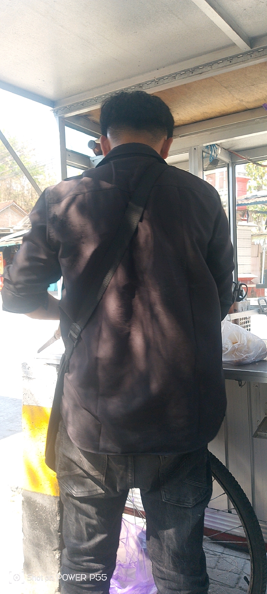 The image, taken in Weru, Indonesia, shows the back of a person wearing a dark collared shirt and dark jeans. The person is standing in front of a food stall, which appears to be made of metal and wood. The stall has a counter with some plastic-wrapped food items and a black bag. A bicycle wheel is visible on the right side of the frame. The background shows a street scene with buildings and trees. The lighting suggests it is daytime.
