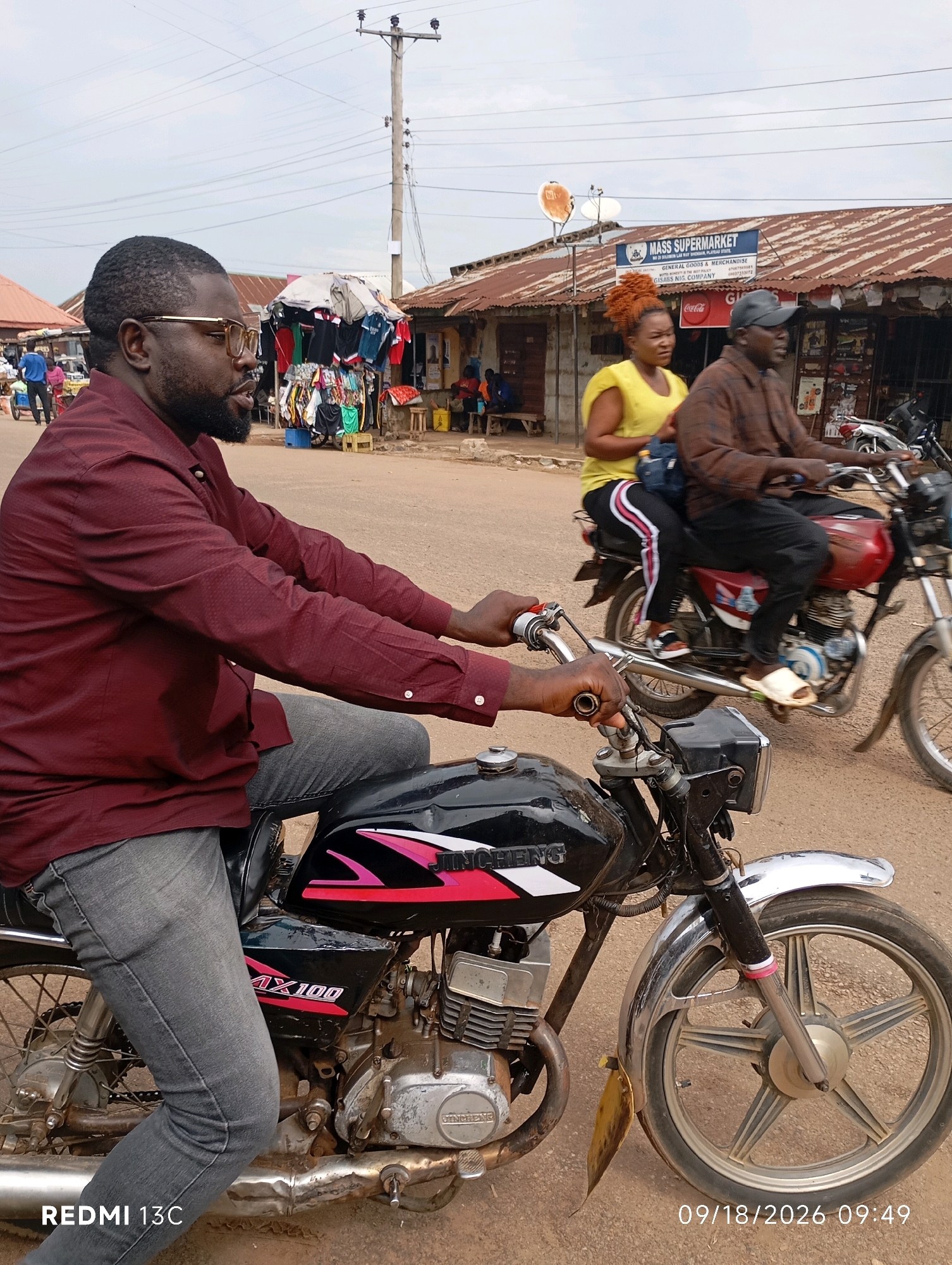 In Shendam, Nigeria, on a dusty road under a cloudy sky, two motorcycles are the primary subjects. The motorcycle in the foreground is black with pink and white graphics and is ridden by a man wearing a maroon long-sleeved shirt, glasses, and grey jeans. His hands are on the handlebars. Behind him, a second motorcycle, predominantly red, carries a man in a brown plaid shirt and a woman in a bright yellow sleeveless top with a blue bag slung over her shoulder. Both riders on the second motorcycle are also looking forward. In the background, stalls with clothing and goods line the road, and a "MASS SUPERMARKET" sign is visible on a building with a corrugated iron roof. Electrical poles and wires crisscross the sky. The timestamp "09/18/2026 09:49" is displayed in the bottom right corner, and "REDMI 13C" is in the bottom left.