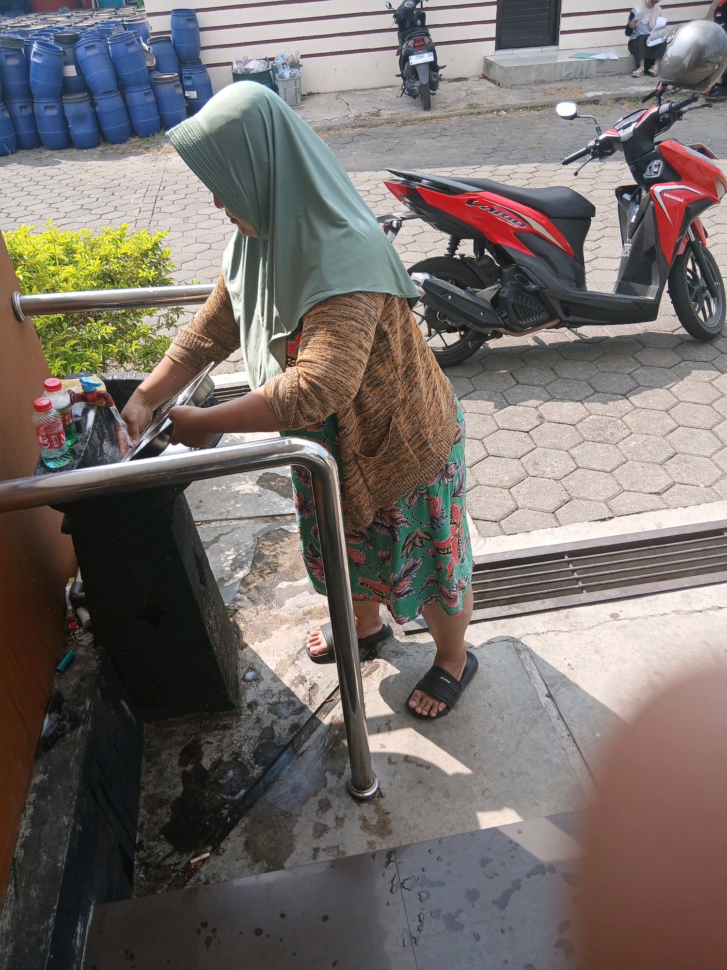 A person wearing a green hijab and a patterned skirt washes a metal bowl under a running faucet at an outdoor sink. Two red-capped bottles, one green and one clear, sit beside the sink. In the background, a red and black Honda motorcycle is parked on a cobblestone surface. Further back, stacks of blue barrels are visible, and two people are seated near a building. The scene appears to be in Jepangpakis, Indonesia.