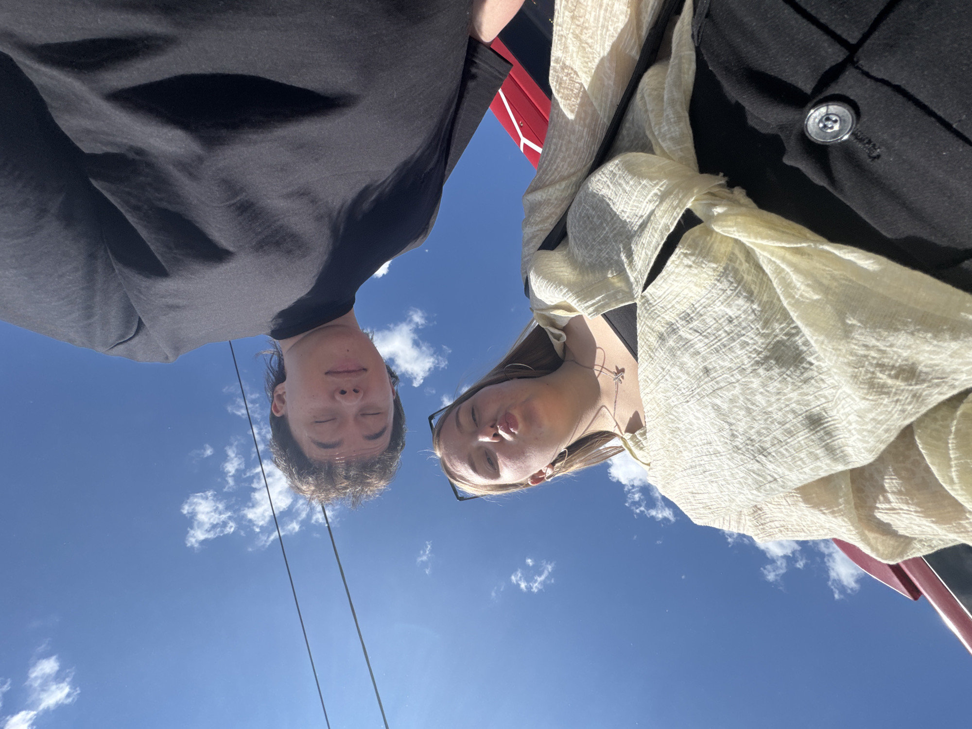 Two individuals, one male and one female, are captured in a low-angle, wide shot against a bright blue sky with scattered white clouds. The perspective is from below, looking up at the subjects. The male is positioned in the upper right quadrant of the frame, with his eyes closed and his head tilted downwards. He is wearing a dark, possibly black, t-shirt. The female is in the lower half of the frame, looking upwards towards the camera with her lips pursed. She has blonde hair, is wearing a light-colored, possibly cream or pale yellow, open-fronted garment over a dark top, and a necklace. She also has sunglasses perched on her head. In the background, a red and black structure, possibly part of a vehicle or a building element, is visible. Two dark lines, possibly wires or cables, run horizontally across the upper portion of the sky. The overall scene suggests an outdoor setting on a sunny day. The location is identified as the city of Perm, Russia, though no specific landmarks within Perm are discernible from the image alone.