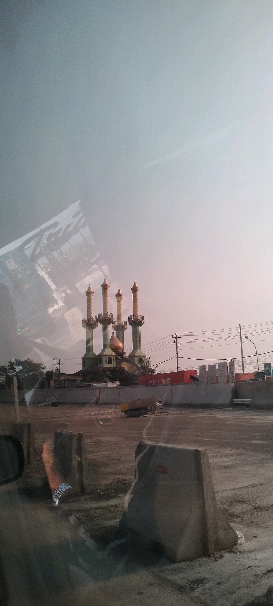 The image shows a mosque with four minarets and a dome, likely under construction or renovation, in Semarang, Indonesia. The mosque is green with gold accents. In front of the mosque, there is a concrete barrier. The foreground of the image appears to be the interior of a vehicle, with the reflection of the exterior visible on the window. The sky is a pale blue with some hazy clouds. The overall scene depicts an urban landscape, possibly a construction site, with a prominent religious building.