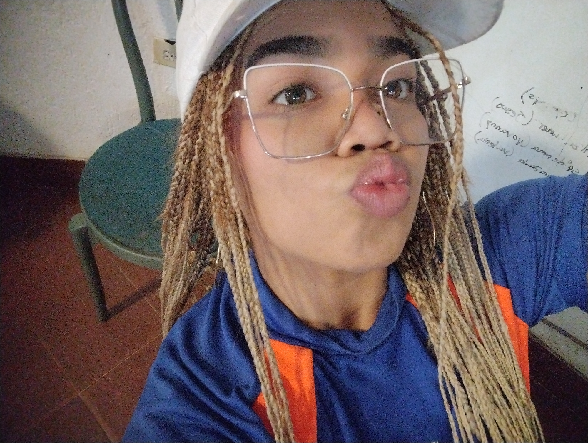 A close-up, slightly tilted selfie features a young person, likely female, captured indoors. The subject has light brown/blonde box braids and wears a white baseball cap, partially visible at the top of the frame. Large, square-framed glasses with light-colored frames sit on their face, through which dark eyes look slightly upwards. The person's lips are pursed in a playful "kiss face" expression. They are wearing a blue long-sleeved top with a prominent orange panel on the front. A gold-colored hoop earring is visible on the right side of the face.
The background shows a simple indoor setting. To the left, a chair with a green/teal seat and a dark frame is partially visible, against a light-colored wall where an electrical outlet is mounted. The floor appears to be covered with reddish-brown tiles. To the right of the subject, a white wall has handwritten text in a dark color. This text is notably mirrored or written in reverse, suggesting the image is a mirror selfie or taken with a front-facing camera mirroring effect. Partially decipherable words and symbols include "(gege)", "(sese)", "(p_q)", "vanner (p_q)", "iq (p_q)", and partial words like "relav" and "rall". The scene conveys a casual and playful mood. The location context is Porlamar, Venezuela.