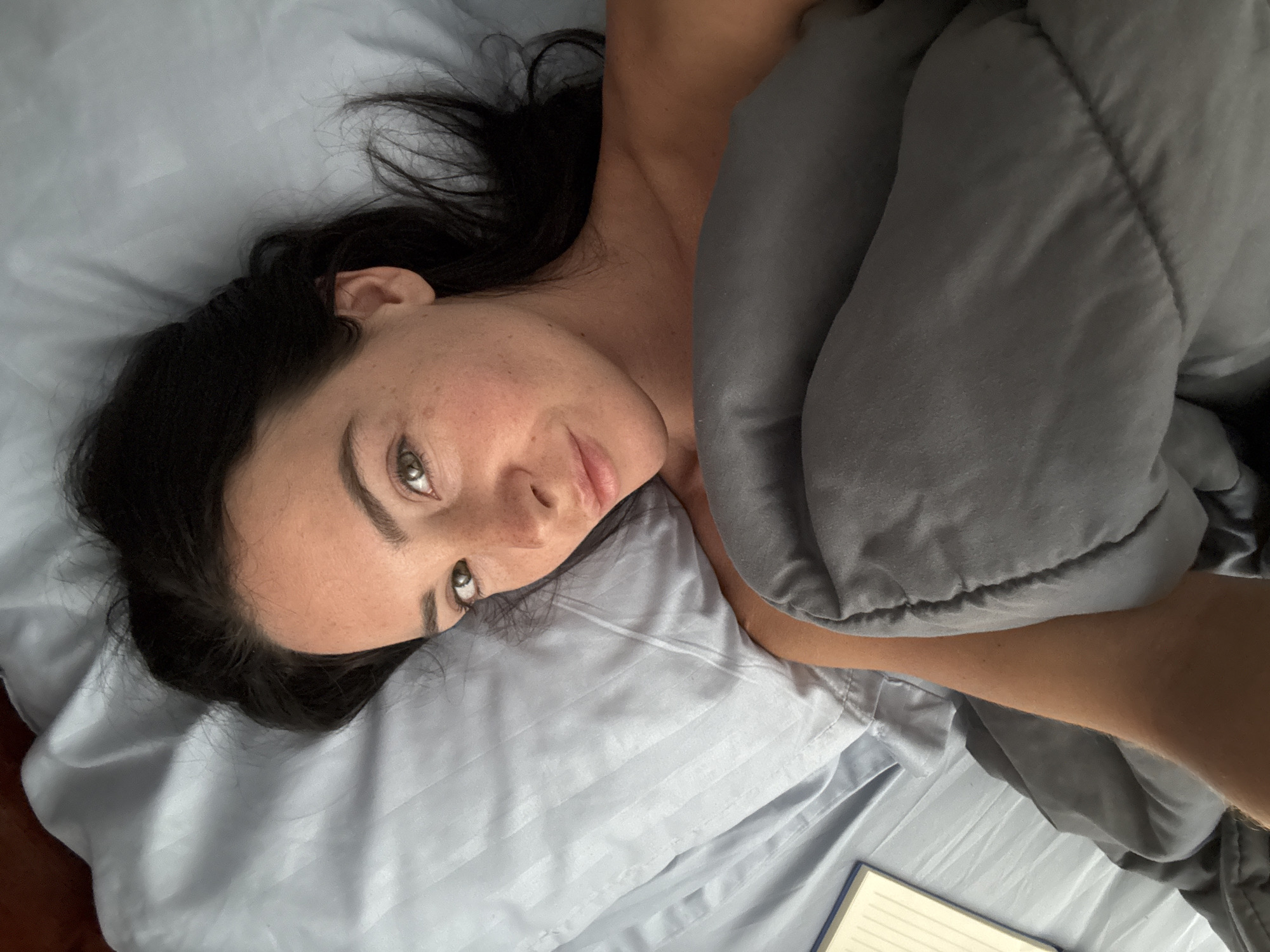A woman with dark hair and green eyes is lying in a bed covered by a grey duvet. She is looking directly at the camera with her head propped up on a light blue pillow. The pillowcase has a striped pattern. A small portion of a notebook with yellow lined pages is visible on the left side of the bed. The environment appears to be a bedroom in North Kuta, Indonesia.
