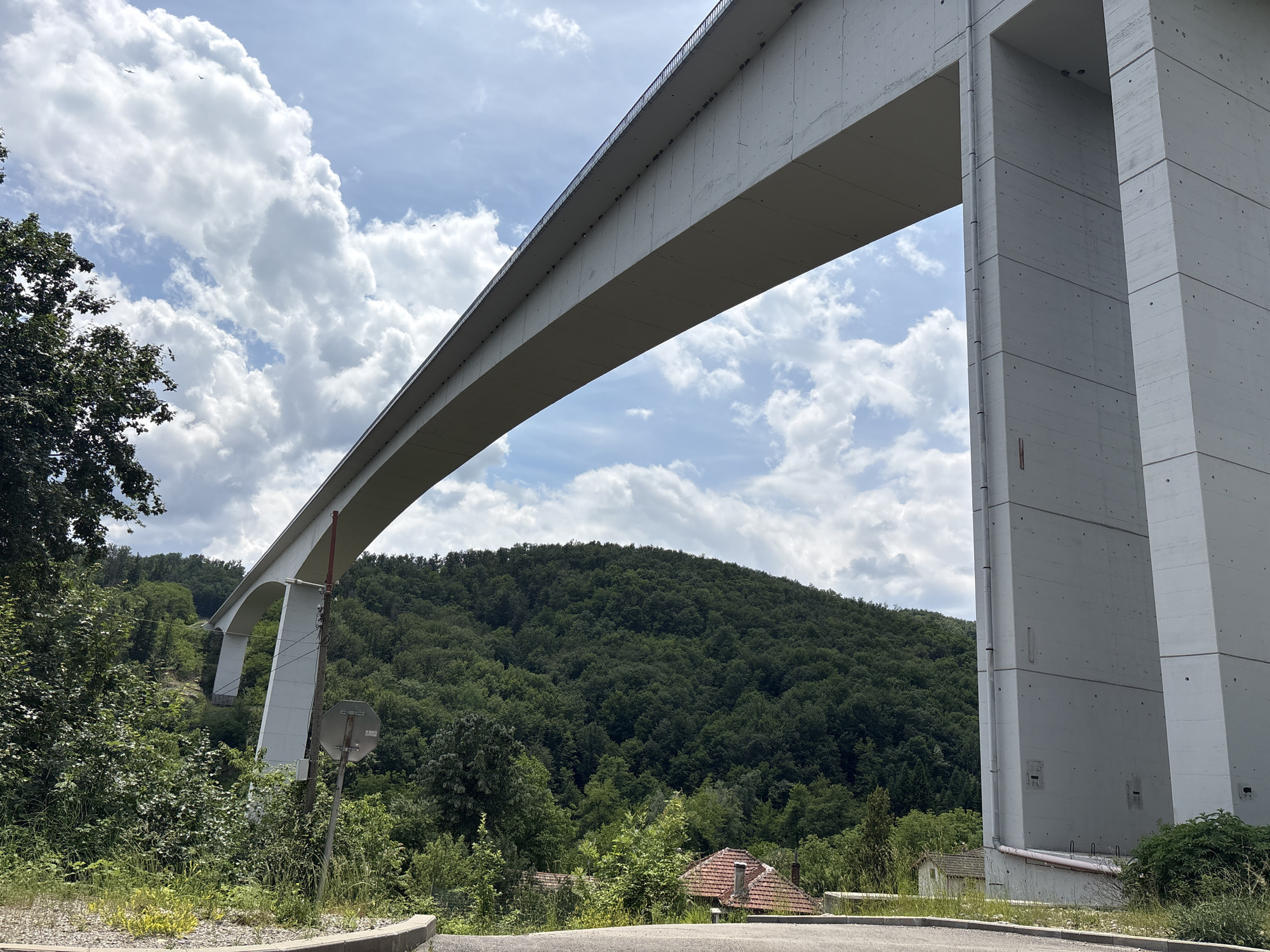 The image shows a large concrete bridge spanning across a valley covered in dense green trees. On the right side of the image, a massive, monolithic concrete pillar supports the bridge. The sky above is a mix of blue and white clouds, with a few small birds visible in the distance. In the foreground, there is a paved road and some overgrown vegetation. A traffic sign is visible on a pole to the left of the bridge pillar. The landscape suggests a rural or semi-rural setting. This bridge is likely the **Vazov Bridge**, also known as the **Gabrovo Viaduct**, located near Gabrovo, Bulgaria.