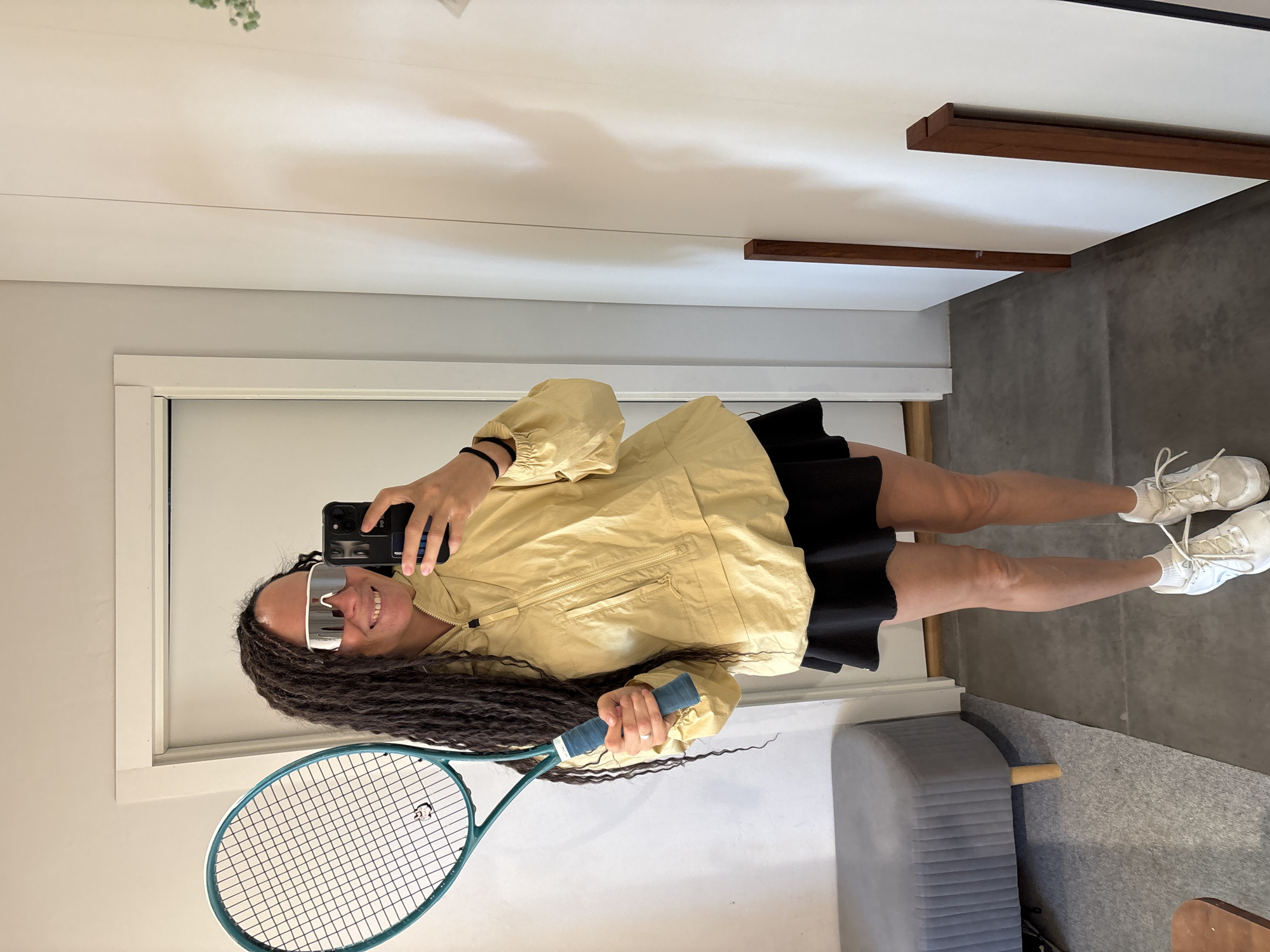 A person stands indoors, holding a tennis racket and a smartphone. They are wearing a light yellow hooded jacket, a black skirt, white socks, and white sneakers. The person also has on large white wrap-around sunglasses. The background shows a white wall, a doorway, and a piece of grey furniture with wooden legs. The floor is covered with grey tiling. The image does not provide any information about the location in the city of Chaikovsky, Russia.