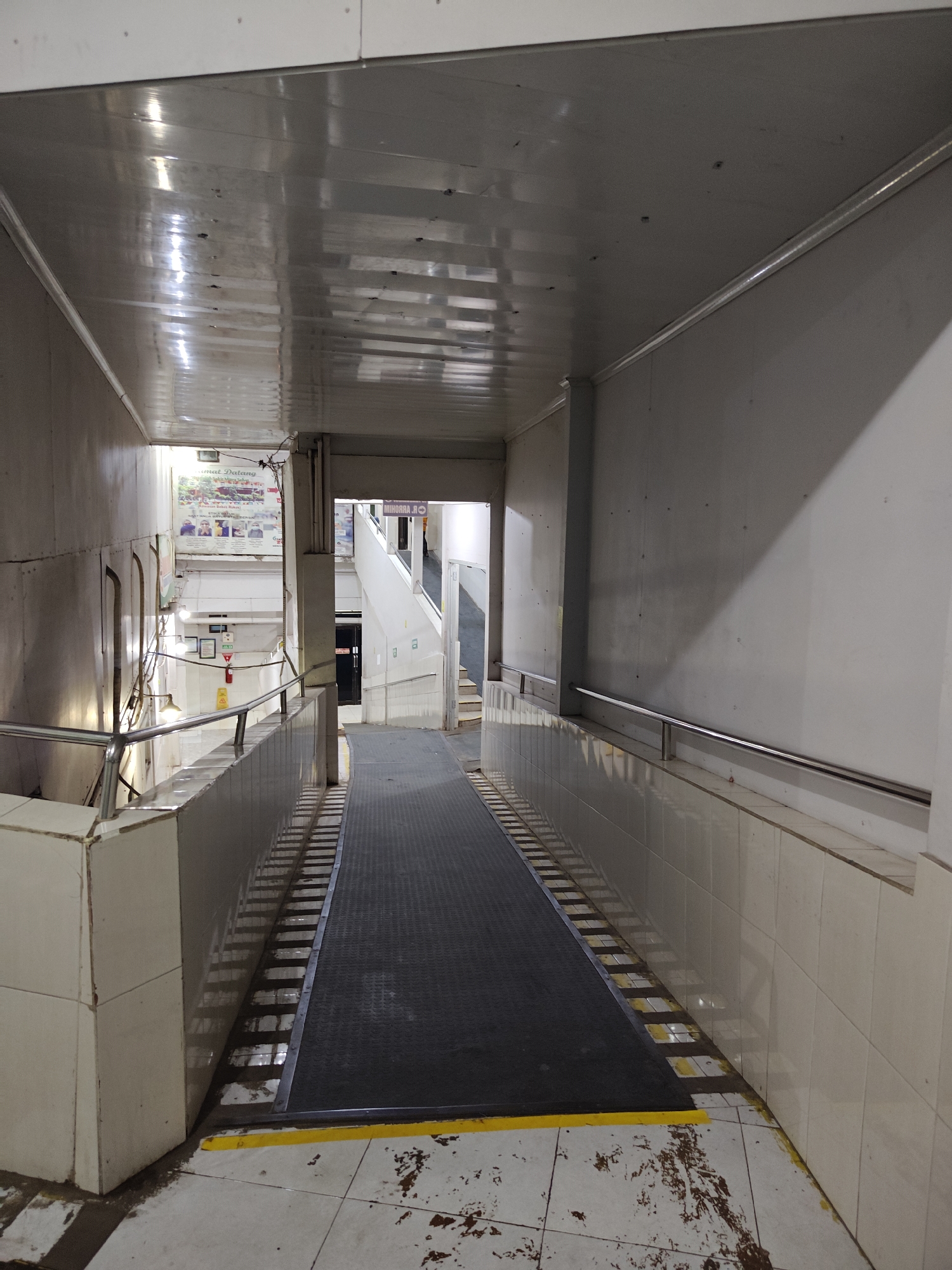The image shows an indoor ramp in a building, likely a public facility or transportation hub, in Situbondo, Indonesia. The ramp is made of dark grey material with yellow markings at the bottom. It is flanked by tiled walls and metal railings. The ceiling is made of reflective white panels, showing some signs of wear and tear. In the background, a staircase is visible, along with a sign that says "SELAMAT DATANG" (Welcome) and other informational posters in Indonesian. The overall environment appears to be functional and somewhat utilitarian, with evidence of foot traffic and use. There are no people visible in the image.
