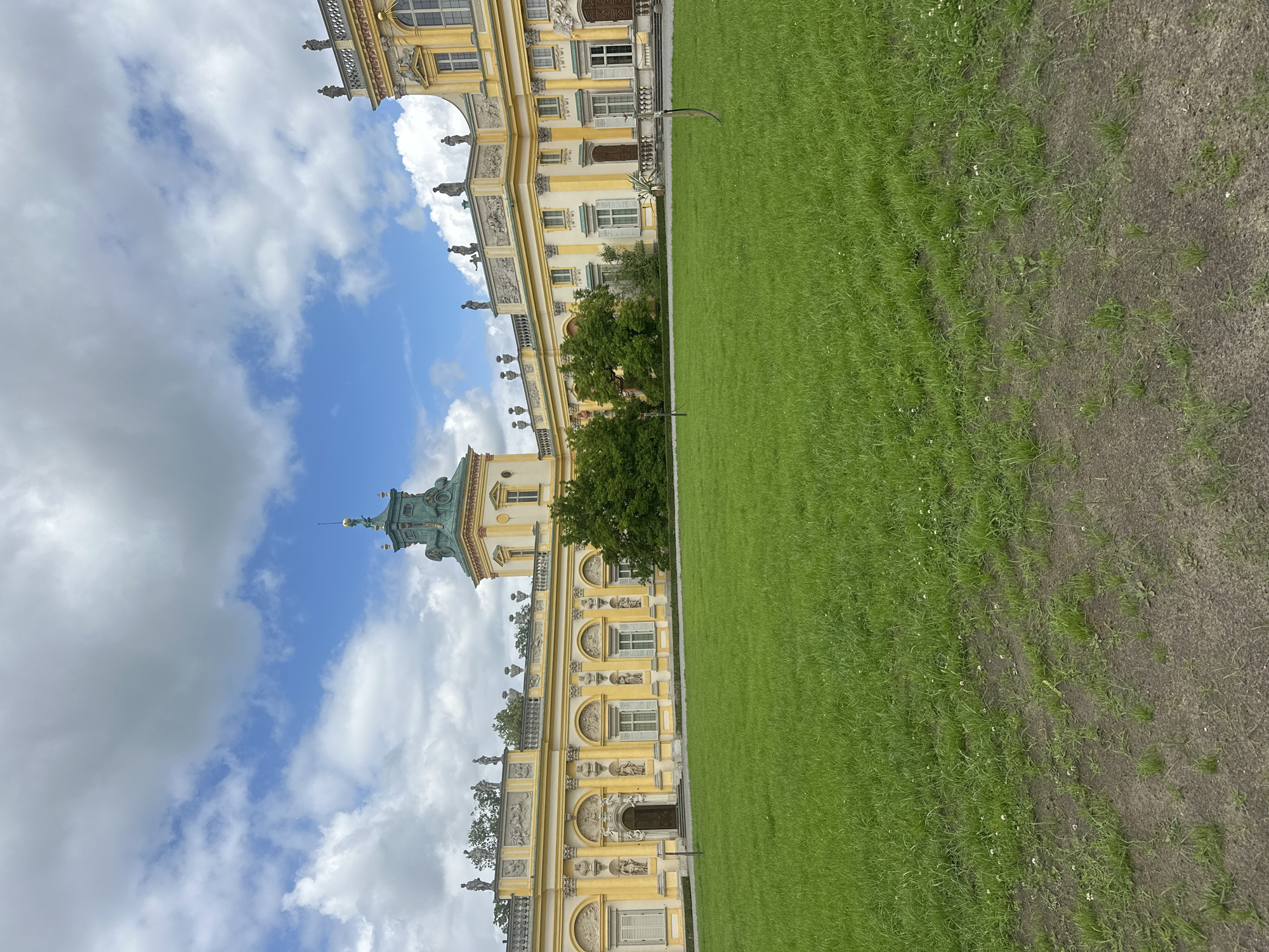 The image shows the facade of a large, yellow, Baroque-style palace with a green-domed tower, set against a cloudy sky. The palace has multiple windows, decorative sculptures, and ornate architectural details. In front of the palace is a large, well-maintained green lawn. The scene appears to be taken during daylight hours. This is the Wilanów Palace in Warsaw, Poland, a historic royal residence. There are no people visible in the image.