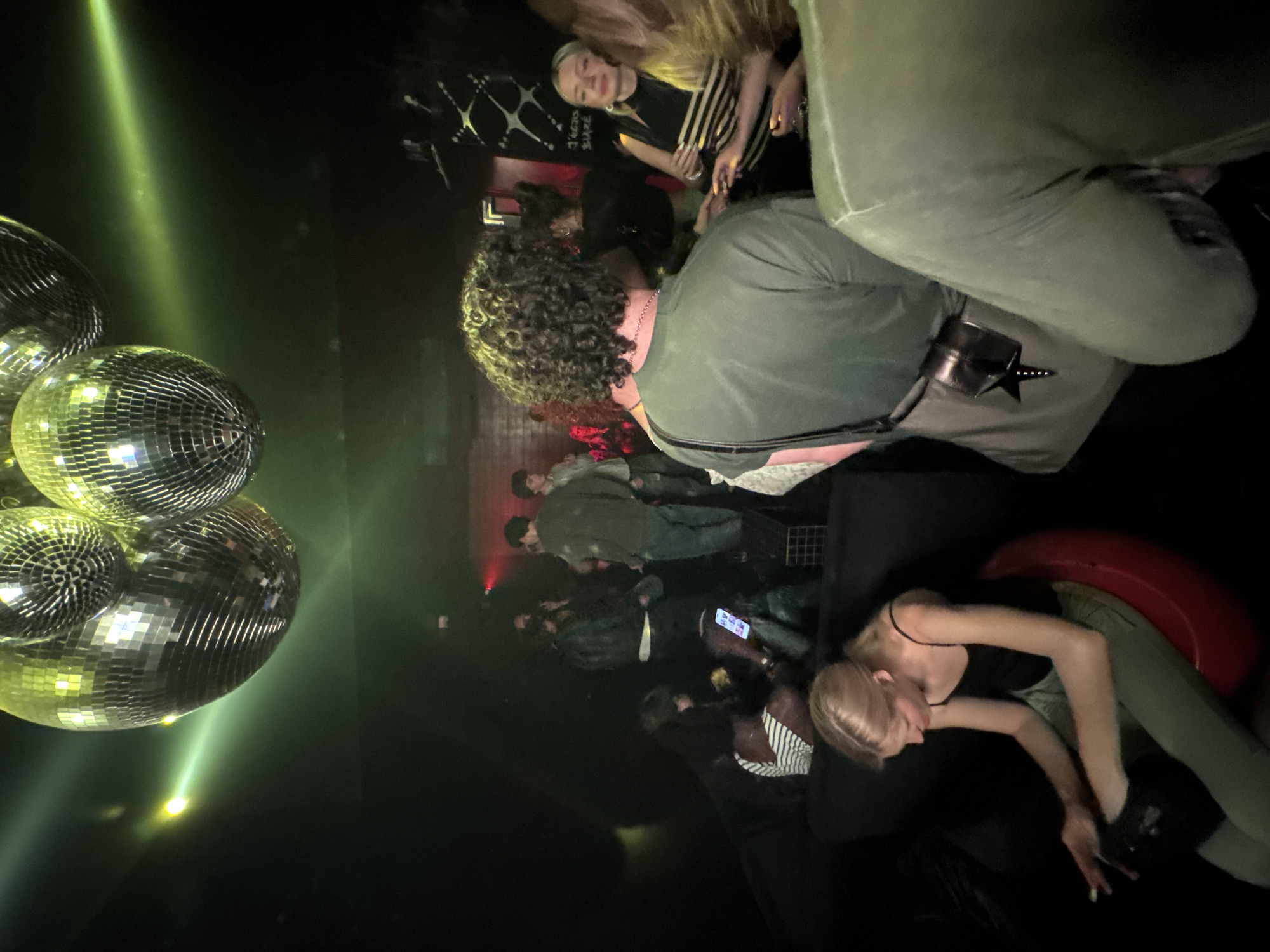 The image depicts an indoor scene, likely a nightclub or bar, with a dark, moody ambiance illuminated by disco balls and green spotlights. Several individuals are present, some standing, others seated. In the foreground, a person with curly hair wearing a green t-shirt is facing away from the camera. To the left, a young woman with blonde hair is seated on a red beanbag chair, looking down at her phone. Other figures are visible in the background, blurred by the low light and movement, suggesting a lively atmosphere. The overall scene captures a moment of social gathering in a dimly lit, party-like environment.
