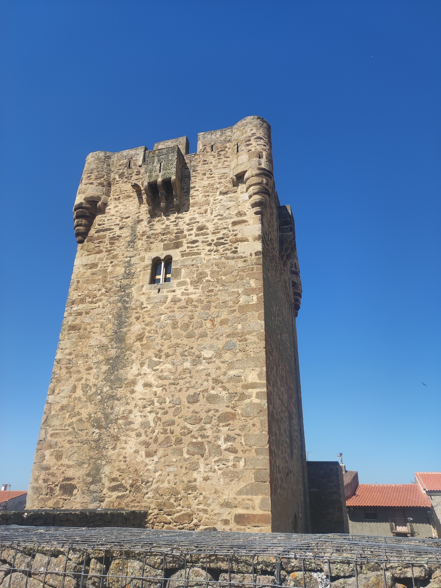 This image captures a detailed view of the Torre del Reloj (Clock Tower) in the city of Sobradillo, Spain, under a clear blue sky. The tower is constructed from rough-hewn, beige stone blocks, showing signs of age and weathering. It features crenellated battlements at the top, with square-shaped openings. Projecting from the upper corners are decorative, circular stone elements. A single, small rectangular window with a simple cross-shaped frame is visible on the main body of the tower.
In the foreground, a low stone wall is partially obscured by a wire mesh structure filled with rocks, which appears to be a retaining wall or a decorative element. Beyond the tower and to the right, the terracotta-tiled roofs of several houses are visible, suggesting a village or town setting. The overall impression is of an ancient, robust structure against a bright, open sky. There are no people or animals visible in the image. The lighting indicates it is daytime.