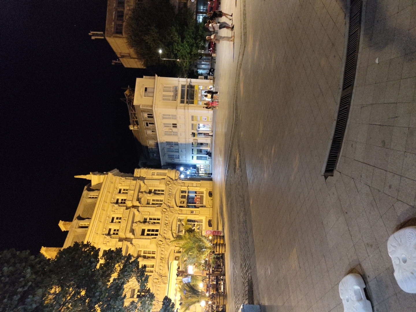 The image depicts a night scene in a city square, likely in Ruse, Bulgaria. The foreground shows a paved area with a drainage grate. In the mid-ground, several buildings are visible. On the left is an ornate, multi-story building with yellow illuminated windows, featuring balconies and decorative architectural elements. Adjacent to it, to the right, is a simpler white building with multiple windows. Further to the right, a larger, more modern building with a lighter facade rises in the background. Several trees are present, including a large one on the left and others closer to the buildings. In the mid-ground and background, small groups of people are visible walking and standing, some appearing to be patrons of an outdoor seating area of a establishment under an awning. The scene is illuminated by artificial lighting from the buildings and streetlights, creating a nocturnal urban atmosphere.