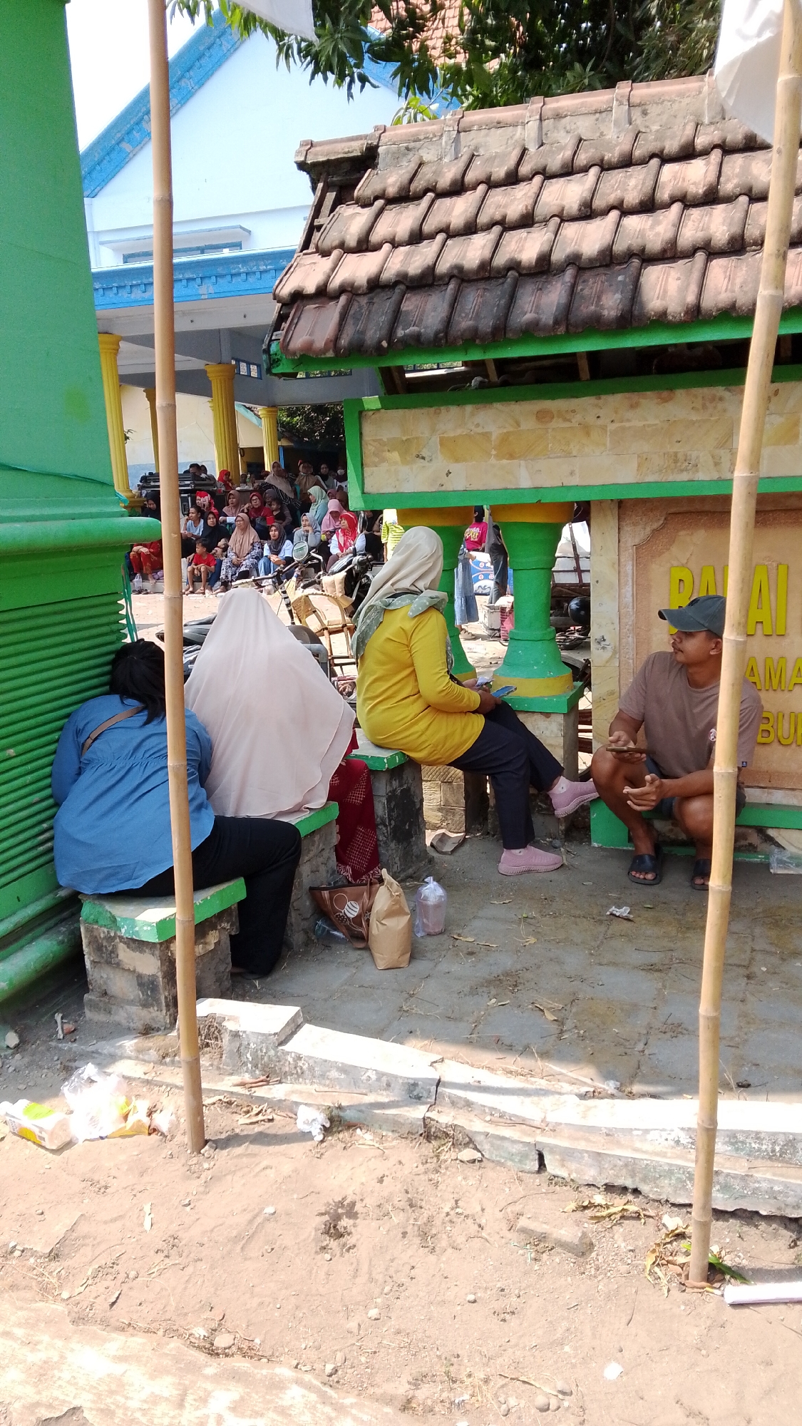 The image captures an outdoor scene in Mancar, Indonesia, likely in a public gathering area or near a building. In the foreground, three individuals are seated. On the left, a person wearing a blue shirt and black pants is seated on a green-edged stone bench, their back to the camera. To their right, another person is seated on the same bench, covered in a light pink hijab and clothing. Further to the right, a woman in a yellow long-sleeved shirt and black pants, also wearing a light-colored hijab, is seated on a similar bench, holding a mobile phone. In the middle ground, a man in a brown t-shirt and dark shorts is crouched down, looking towards the woman in yellow, and also holding a mobile phone. Behind them, a crowd of people are visible, some seated and some standing, with a motorcycle parked amongst them. The environment features a building with a tiled roof and visible pillars, as well as a green structure in the foreground. The ground is sandy and dirt, with some scattered debris. A bamboo pole stands vertically in the foreground, partially obscuring the scene. The overall impression is of a casual gathering or waiting area.