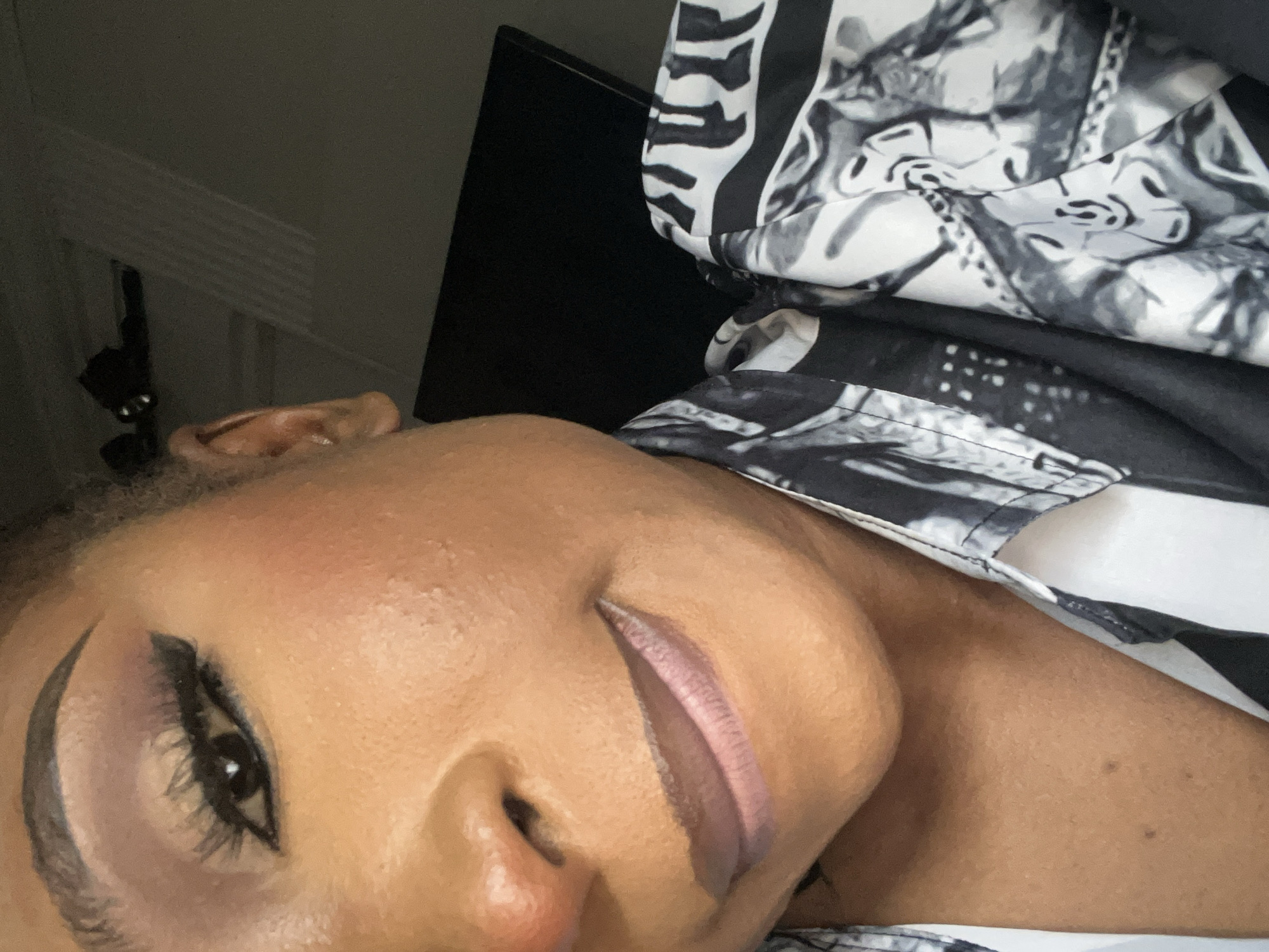 The image is a close-up, eye-level, slightly tilted selfie of a Black woman. She is looking towards the viewer, with her head angled to her right. Her left eye is in focus, showcasing brown eyeshadow with a winged eyeliner and long, dark false eyelashes. Her skin has a warm, brown tone with visible texture. She is wearing a light pink lipstick with a matte finish. The background is dark and out of focus, with a hint of a black monitor or screen to the right and what appears to be an ear visible on the far left. The woman is wearing a black and white patterned shirt. The pattern on the shirt appears to be abstract or graphic. There are no discernable landmarks of Abuja, Nigeria visible in this close-up image.