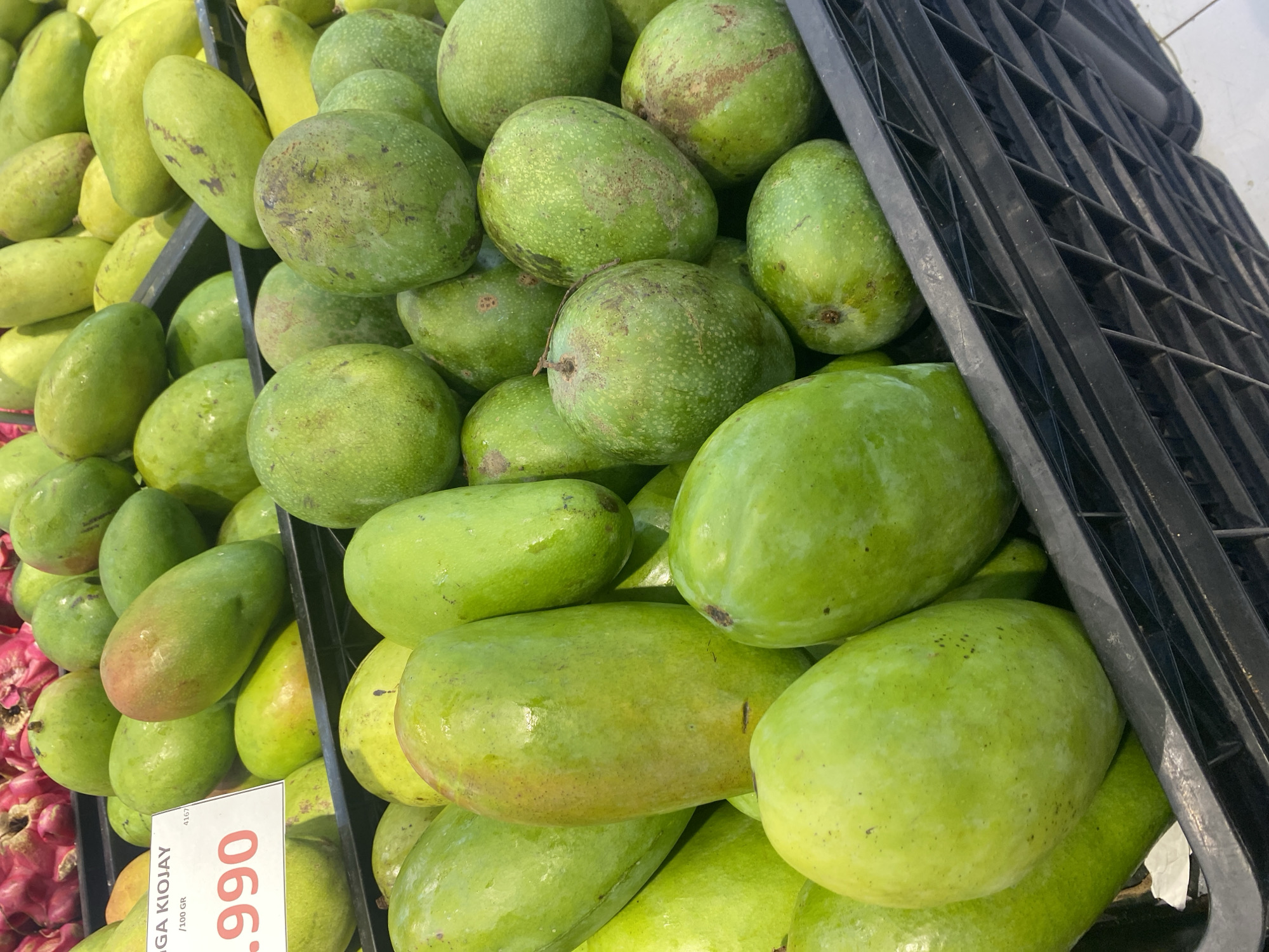 A close-up, slightly angled shot shows a black plastic crate filled with green mangoes. A small white price tag with red text is visible in the upper left corner, reading "GA KIOJAY .990". The background shows more green mangoes and some red dragon fruit in the upper left. The setting appears to be a market stall in Watampone, Indonesia.