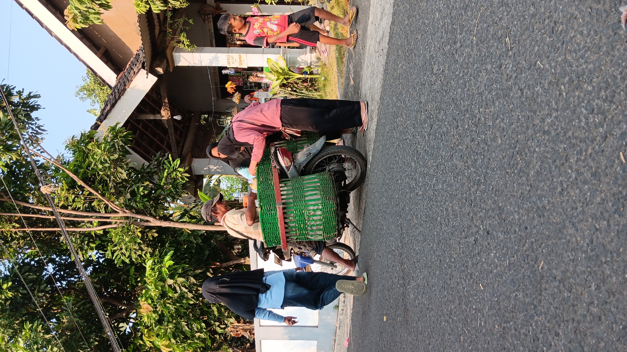 In Cerme, Indonesia, on a street with a paved asphalt surface, a group of people are gathered around a motorcycle with a large, green woven basket mounted on the back. A man wearing a cap and a light-colored shirt is seated on the motorcycle, interacting with two women. One woman, dressed in a pink top and black pants, leans towards the basket, and another woman in a black hijab stands behind her. To the left, a person wearing a blue shirt and dark pants walks away from the motorcycle, their back to the camera. To the right, a young boy in a red and black t-shirt and shorts walks along the side of the road. Behind the group, a building with a tiled roof and white pillars is visible, with trees and vegetation surrounding it. The scene appears to be in a residential or commercial area, with the sky visible in the background.