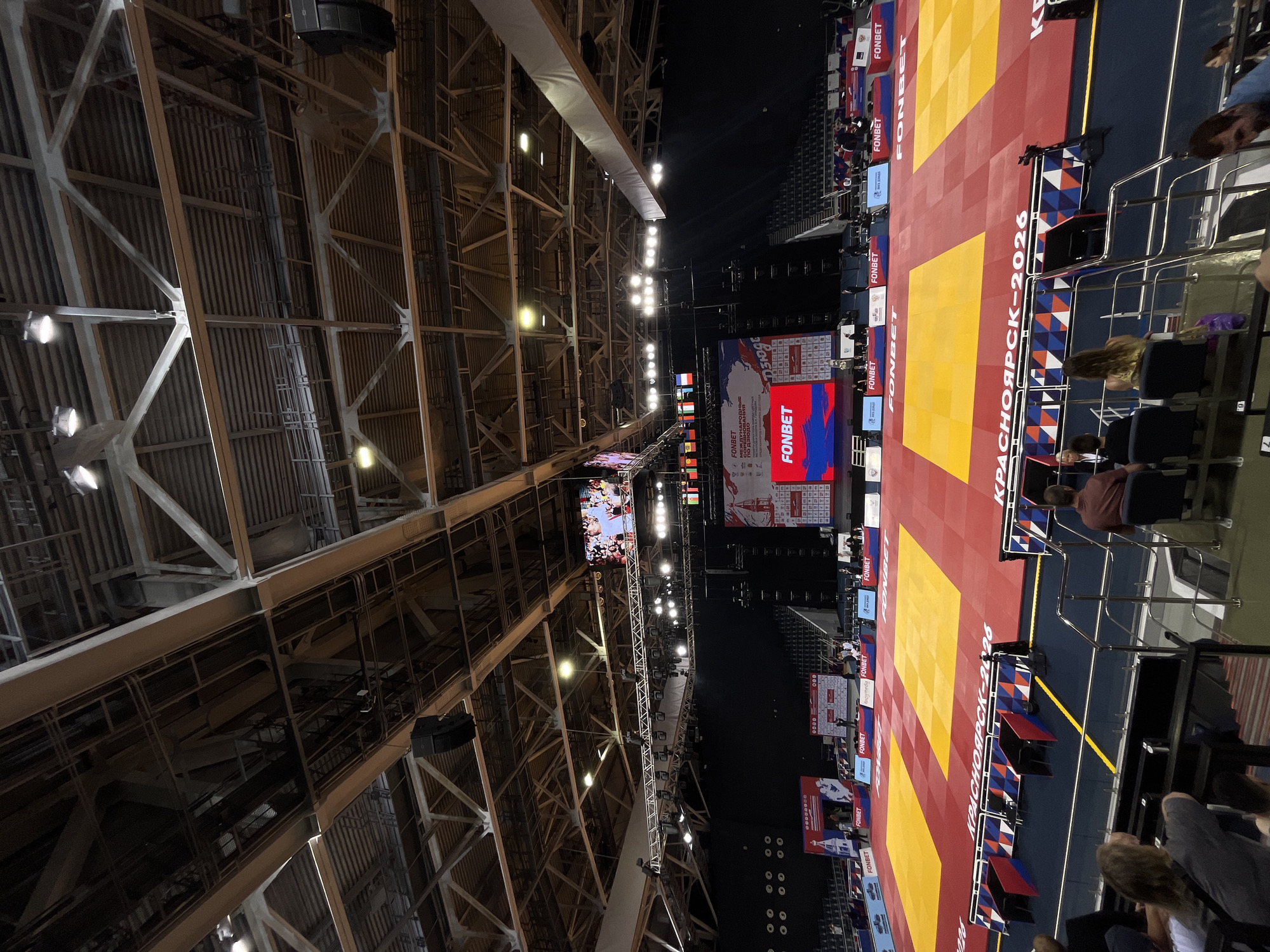 An indoor sporting arena is captured in a wide shot, likely within Krasnoyarsk, Russia, given the prominent text "Krasnoyarsk-2026" visible on the matting. The arena features a large, brightly lit competition mat with alternating red and yellow sections, designed for a sport like judo or wrestling. Spectator seating, partially occupied by a few individuals, is visible in the foreground and receding into the background. Above the competition area, a large video screen displays event information and sponsor logos, including "FONBET." The ceiling is a complex structure of industrial beams, trusses, and lighting rigs, characteristic of large sports venues. Several large speakers are suspended from the ceiling. The overall atmosphere suggests an ongoing or recently concluded sporting event.