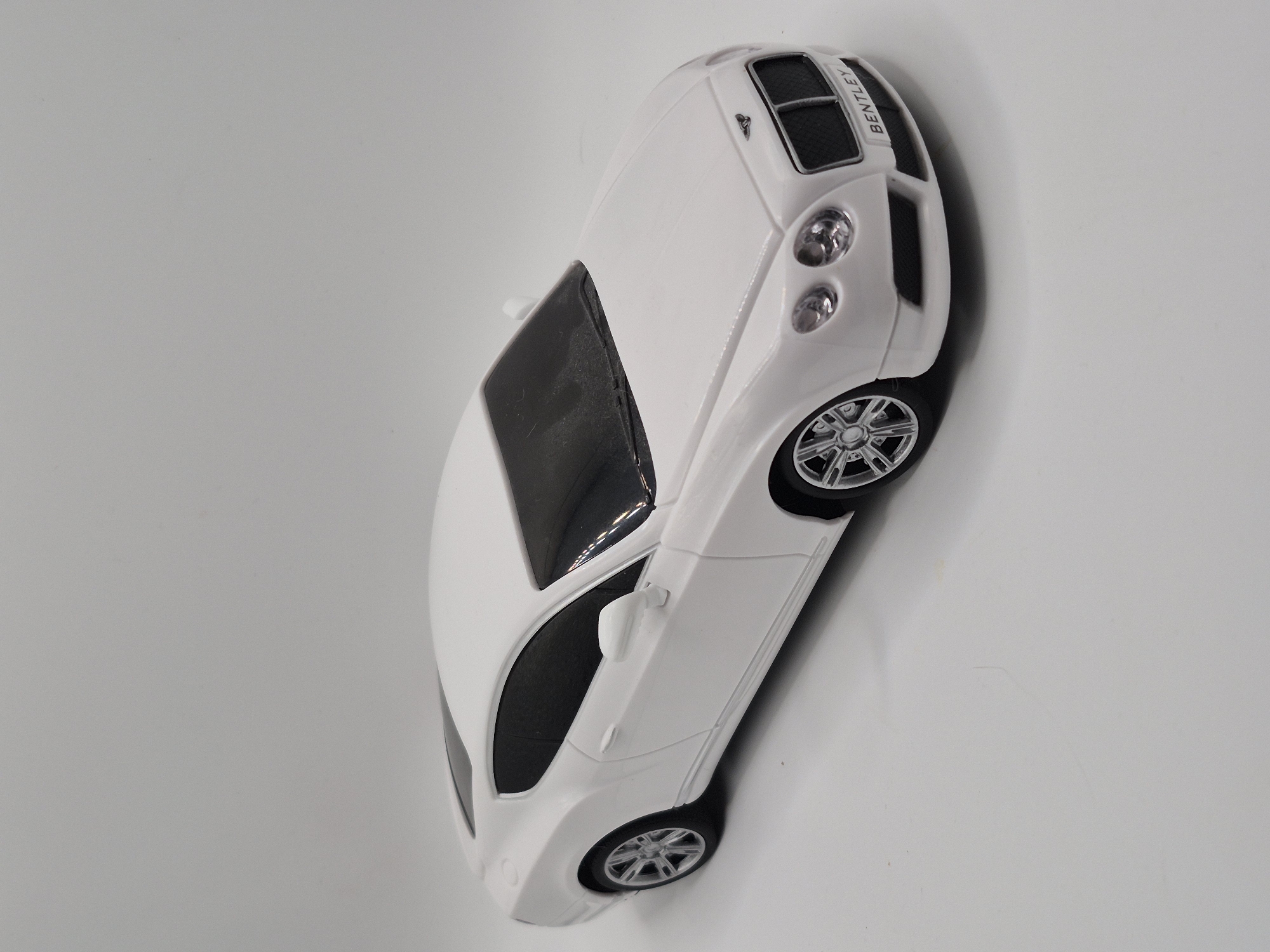 A white toy Bentley Continental GT is photographed against a plain gray background. The car is positioned at an angle, with its front facing towards the right of the frame. The toy car is detailed, featuring realistic headlights, a grille with the Bentley logo, and wheels. The word "BENTLEY" is visible on a license plate at the front of the car. The lighting is even, casting a subtle shadow on the ground beneath the car. The image does not contain any information about the city of Schwäbisch Gmünd, Germany, nor does it depict any people or specific actions.