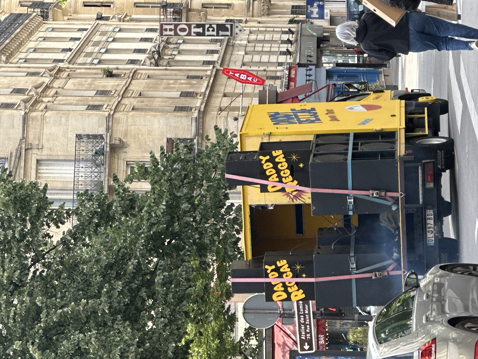 The image depicts a scene in Paris, France, featuring a yellow truck with "DADDY REGGAE" painted on its sides. The truck is equipped with large speakers and is parked on a street lined with buildings. The sign reads "Atelier des Lumières, 38, Rue Saint Maur". A person is walking in the background carrying a box. Other details include a "HOTEL" sign and a red "ABAC" sign on the buildings in the background. A car is parked next to the truck.