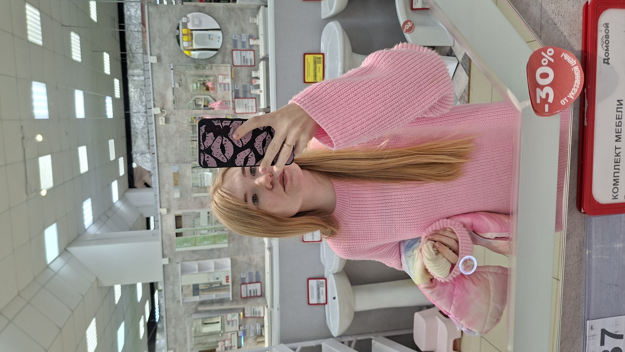 A young woman with long blonde hair and wearing a pink sweater is taking a selfie in a mirror in what appears to be a home furnishings store in Chaikovsky, Russia. She is holding a black phone with a pink lips design on the case. Behind her, several sinks and cabinets can be seen on display, along with signage in Russian and a "-30%" discount sign. The background also shows a wide-angle view of the store's interior with bright overhead lighting and various displays. The overall impression is a casual, self-portrait taken in a commercial setting.