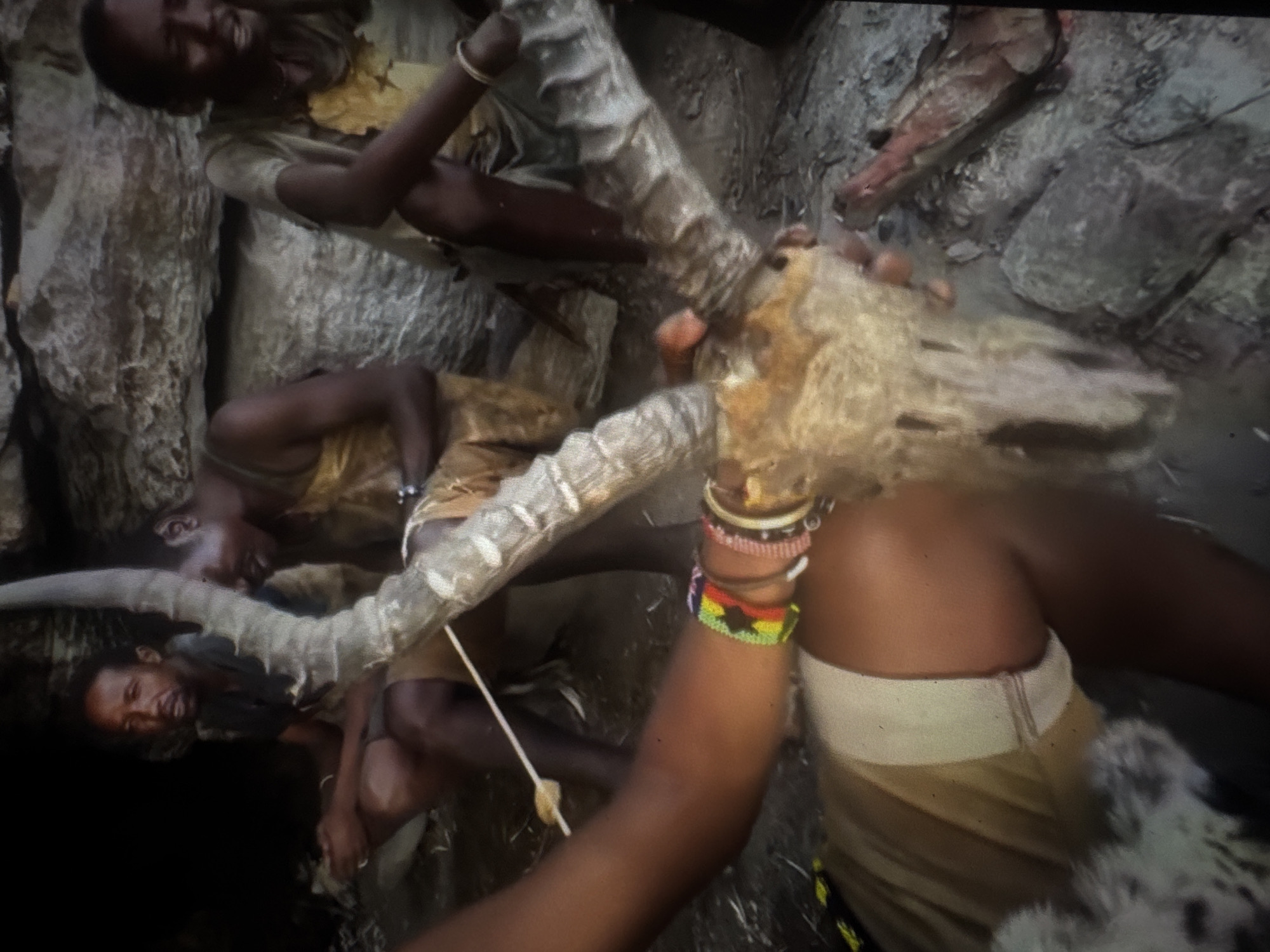 The image depicts a group of people gathered in what appears to be a cave or rock shelter, with a rocky, uneven ground surface. There are at least three individuals visible, dressed in simple, earth-toned garments, possibly made of animal hides or coarse fabric. One person in the foreground is holding a large animal skull with prominent horns. Their arm is adorned with colorful beaded bracelets. Another individual is seated to the left, looking towards the camera with a somber expression. A third person is seated to the right, partially obscured, with their head bowed. The environment suggests a natural, possibly remote setting, with rough rock formations forming the background. There are no clear indications of an urban environment or specific landmarks that would place this scene within the city of Abuja, Nigeria. The overall atmosphere is one of tradition or ritualistic activity.