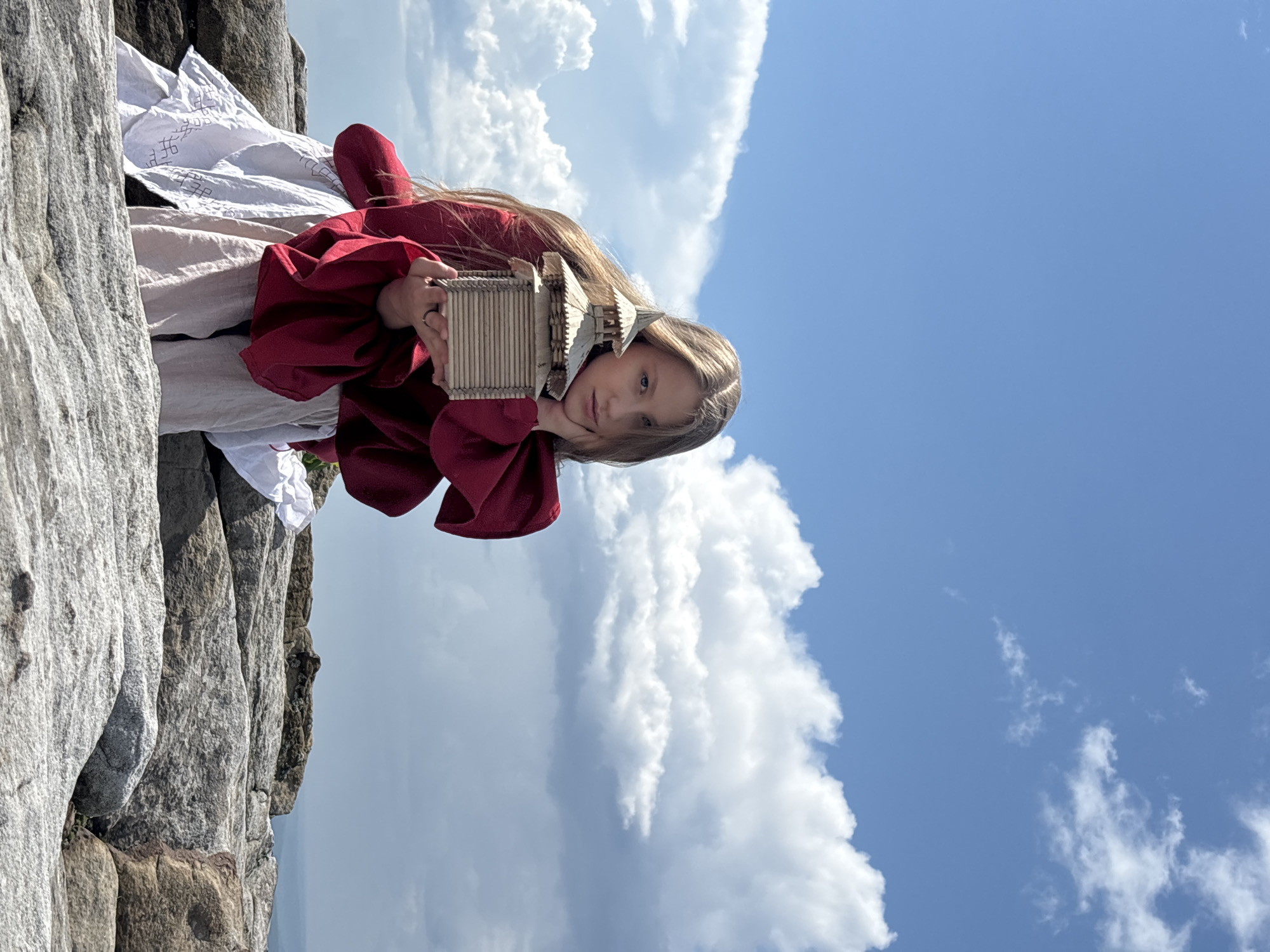 A young Caucasian female with long blonde hair is sitting on a rocky outcrop, holding a miniature wooden house. She is wearing a red top with full sleeves and a white skirt. Her left hand is supporting the wooden house, while her right hand is resting on her cheek. The background consists of a blue sky with white clouds and a distant landscape. The scene appears to be set in a natural, outdoor environment, possibly on a mountain or high plateau. The location is identified as Gubakha, Russia.