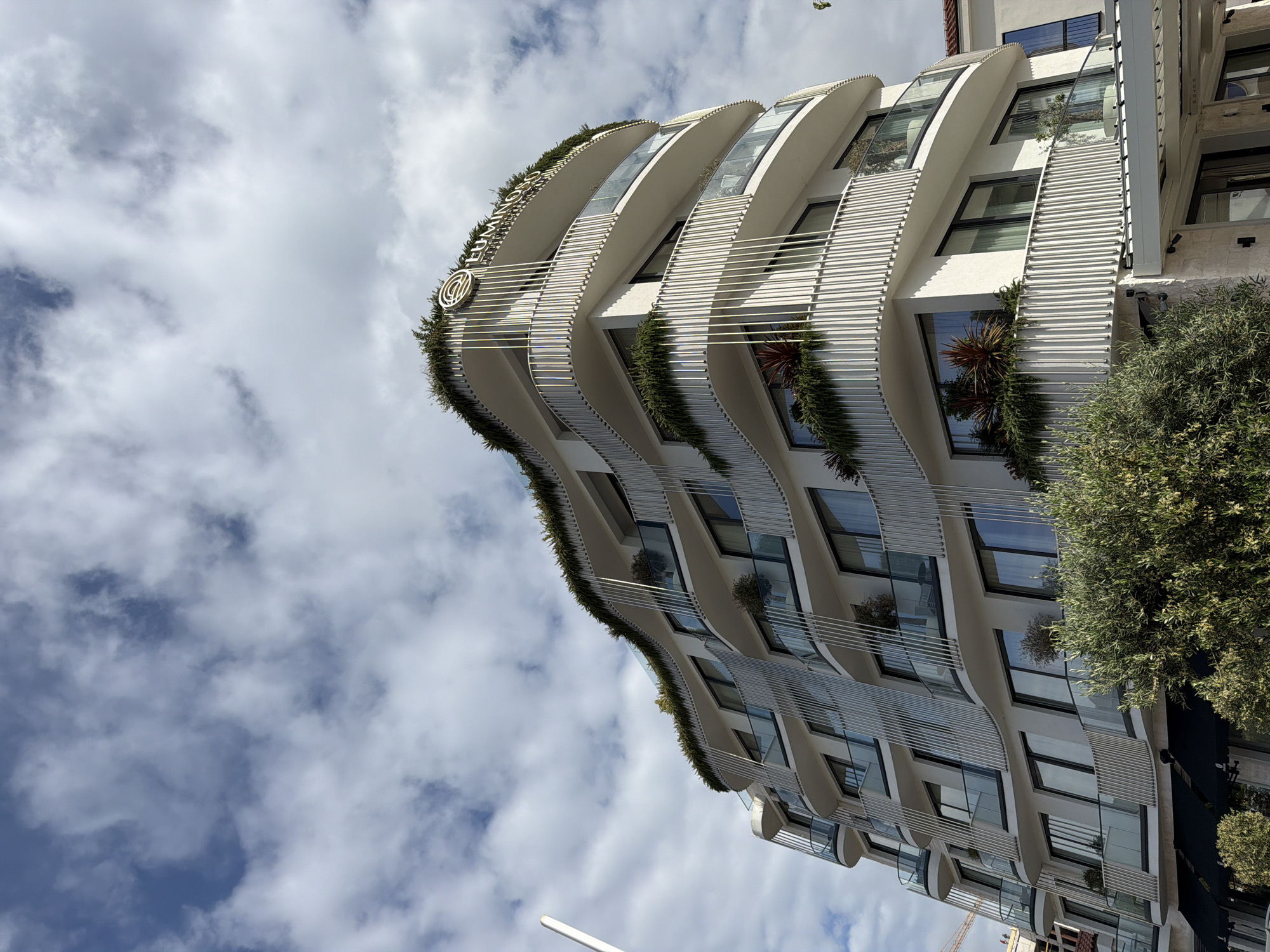 A low-angle, medium shot depicts a modern, multi-story building in Budva, Montenegro, set against a cloudy sky. The building's facade is characterized by curved balconies adorned with greenery. The balconies feature horizontal metal railings and potted plants, creating a terraced garden effect. Large windows reflect the sky. The building is white. A round logo is visible on the building. A tree is to the right of the building. A construction crane is visible in the lower right corner. The orientation of the image is rotated, presenting the building at an angle.