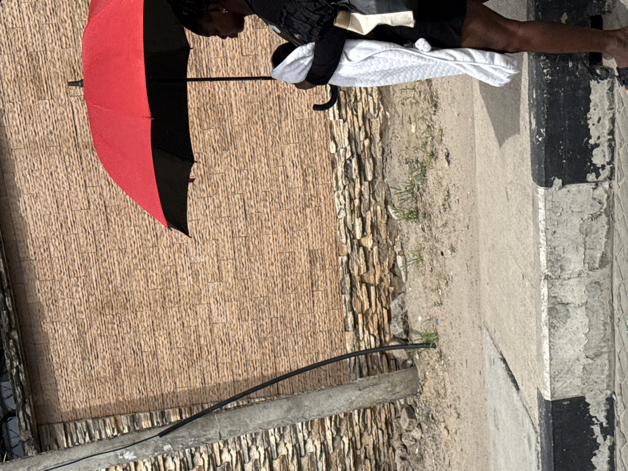 A woman stands on a sidewalk next to a wall holding a baby wrapped in a white blanket while holding a red and black umbrella. The location appears to be Itirin, Nigeria.