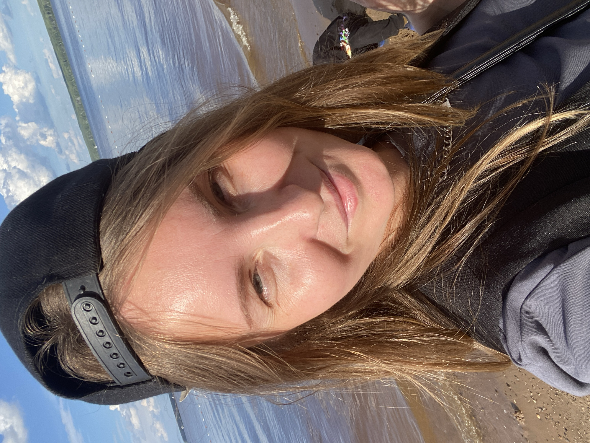 A young woman wearing a black baseball cap and a grey long-sleeved shirt is taking a selfie on a beach. She has long blonde hair and is smiling. The background shows a body of water, likely the Kama River in Chaikovsky, Russia, with a blue sky and clouds above. The beach is sandy and there are some small waves lapping at the shore. There is another person in the background, partially obscured.