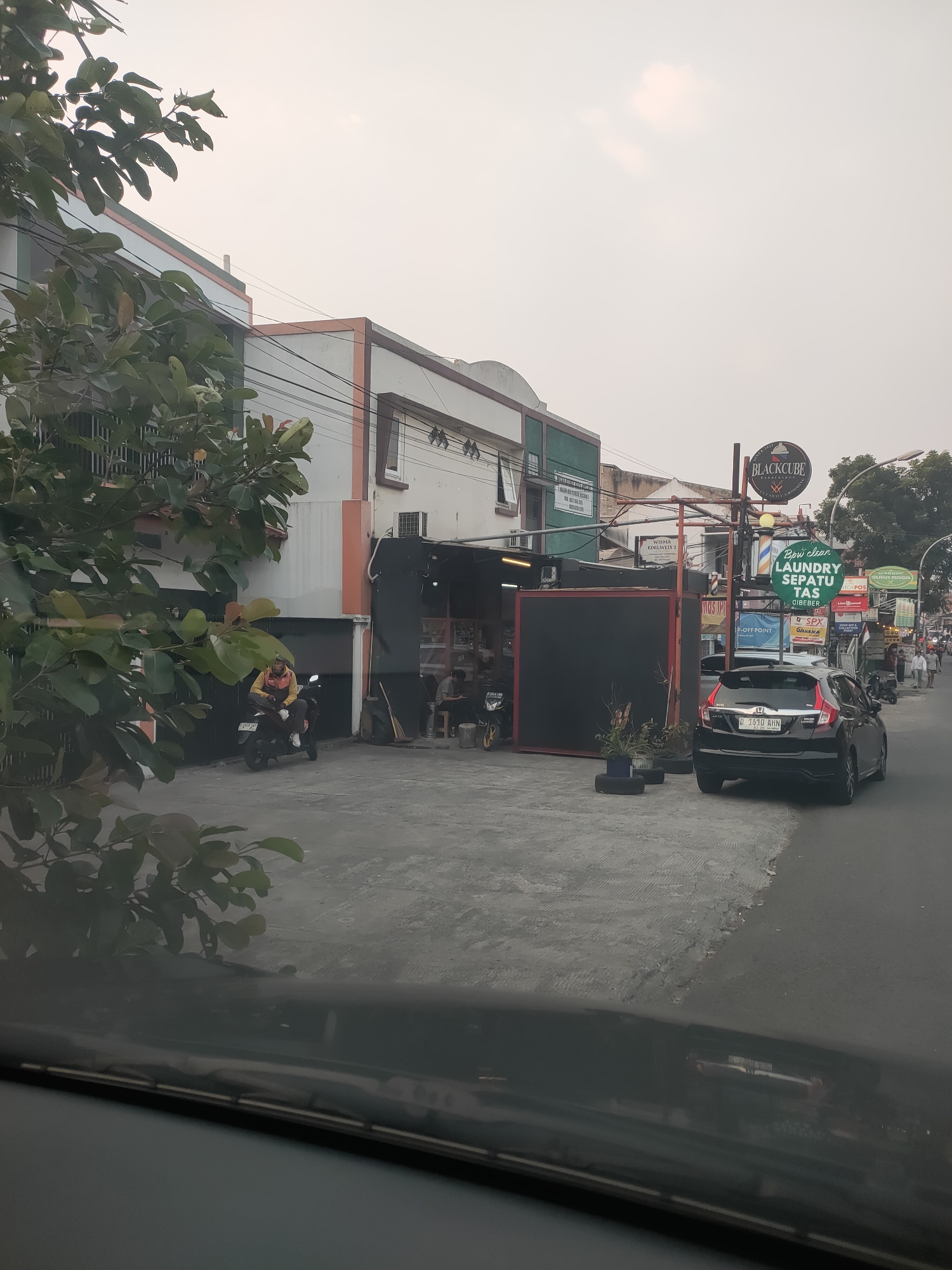 The image captures a street scene in Cimahi, Indonesia, likely in the afternoon given the soft, diffused light. In the foreground, a dark-colored car is parked on the right side of the street, facing away from the viewer. The car is a Honda, with license plate D 1610 AHN. To the left of the car, a paved area appears to be an entrance to a building. A motorcycle with a rider wearing a yellow and orange jacket is parked on this paved area, facing left.
The buildings lining the street are commercial establishments. On the left, a multi-story building with white, orange, and green accents is visible. Trees with lush green foliage partially obscure the view of this building. Further down the street, a structure with a black facade has an open entrance, with several people seated inside or nearby. Above the open entrance, a circular sign with the text "BLACKCUBE BARBERSHOP" is prominently displayed. Another sign, green in color, reads "LAUNDRY SEPATU TAS CIBER". Various other signs for businesses are also visible, indicating a commercial district. The street itself is asphalt, and there are power lines running overhead. The overall impression is one of a busy urban environment with active commerce.