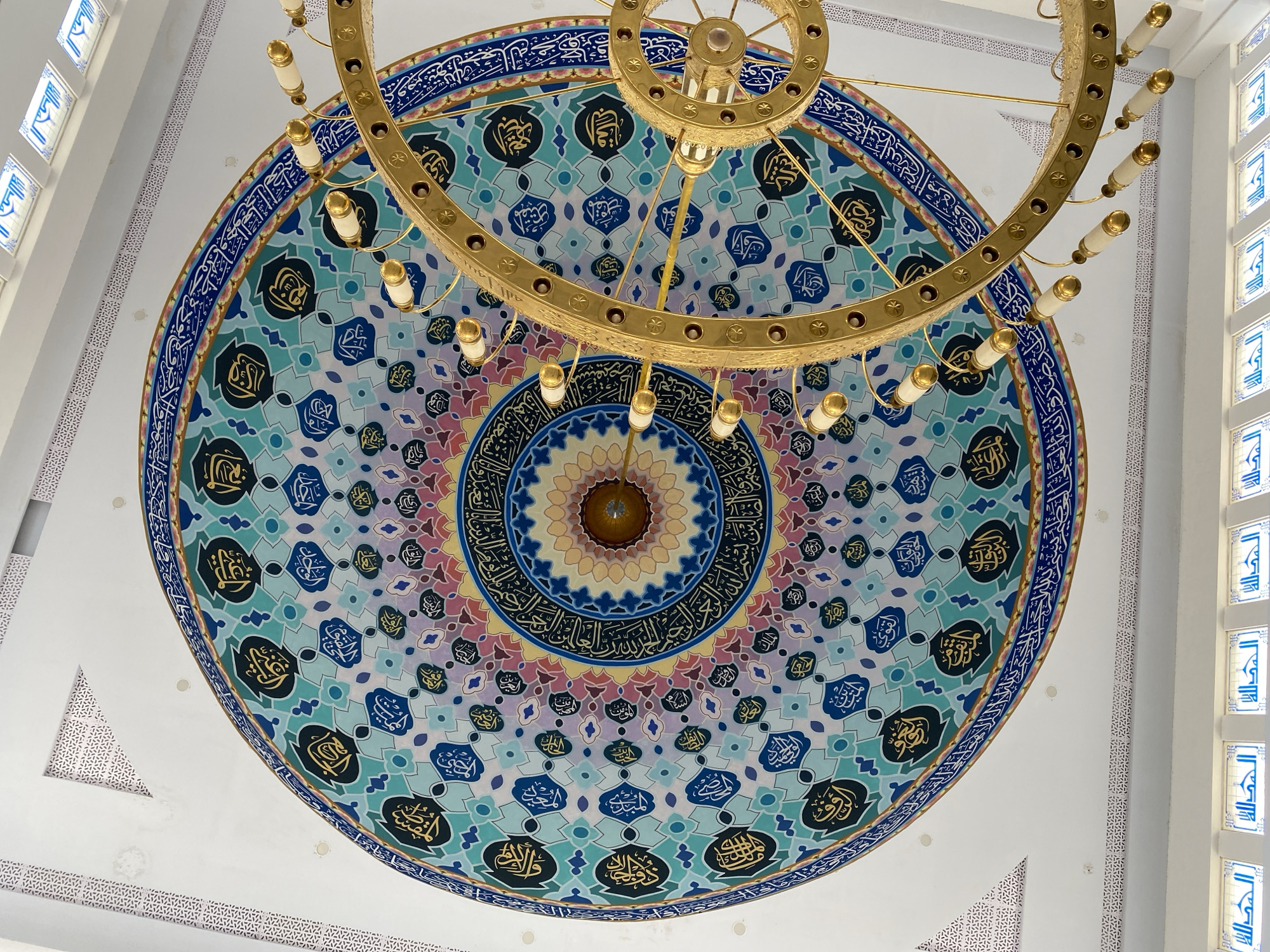 The image shows the interior of a mosque in Lhokseumawe, Indonesia, with a focus on the ornate ceiling and a large, golden chandelier. The ceiling is dominated by a colorful dome decorated with Islamic calligraphy and intricate geometric patterns in shades of blue, green, purple, and pink. The calligraphy is in Arabic script and appears to be religious verses. A large, multi-tiered golden chandelier with white cylindrical lights hangs prominently in the foreground, partially obscuring the central part of the dome. The surrounding ceiling is white, with decorative borders featuring a repeating geometric motif. Below the dome, the image shows several rectangular windows with blue calligraphy on a white background. The overall impression is one of grandeur and religious artistry.