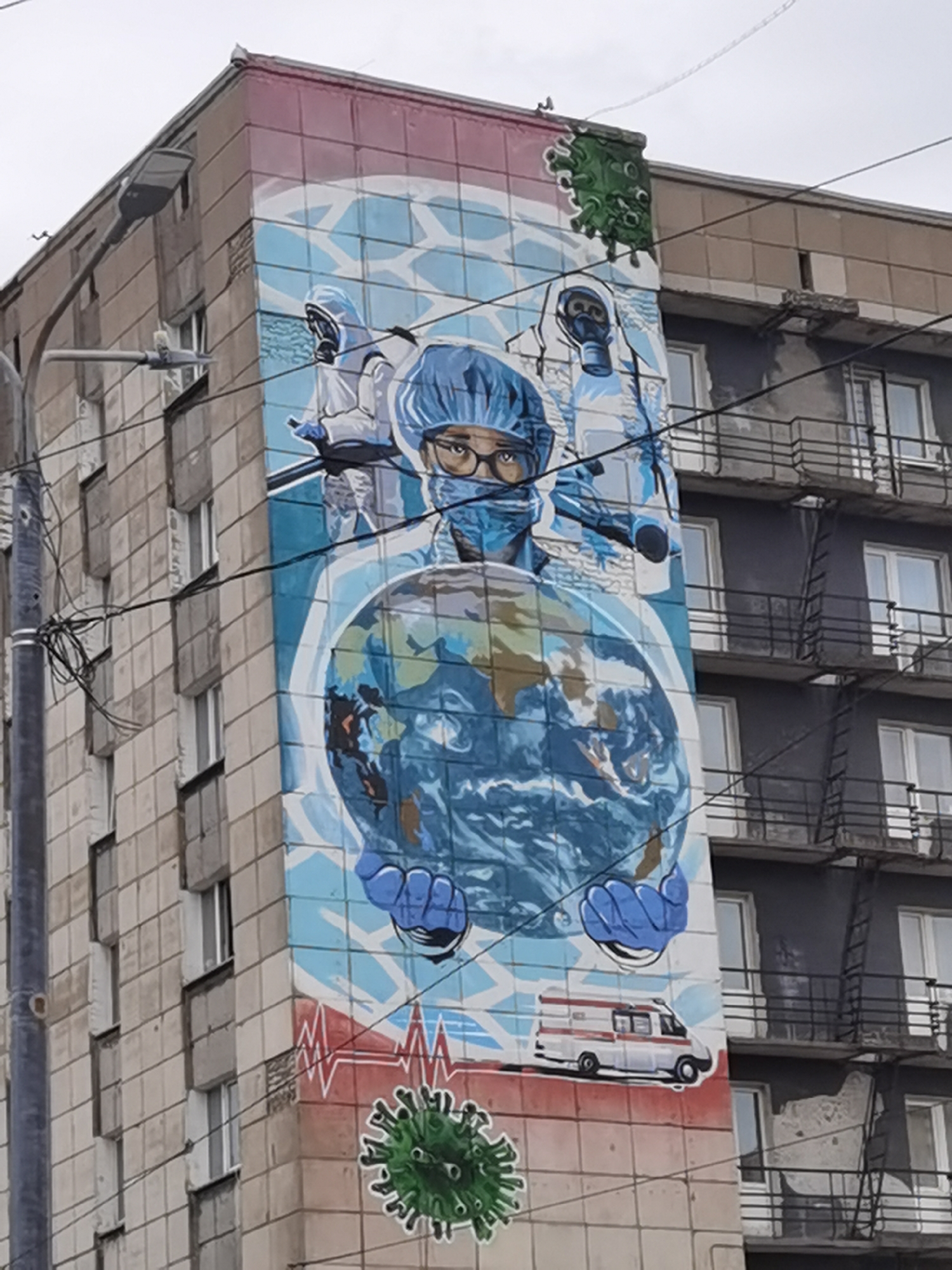 An eye-level, medium-shot photograph shows a mural painted on the side of an apartment building in Perm, Russia. The mural is dominated by a depiction of medical personnel in protective gear, holding a globe. A person in a blue surgical cap and mask, with glasses, is centered, with their gloved hands holding the globe. Behind them are two figures in full protective suits, including respirators. Above them, a green coronavirus particle is painted, with a similar one near the bottom of the mural. An ambulance is also painted below the globe, and a red heartbeat-like line runs horizontally near the bottom of the mural. The mural is painted on a section of the apartment building that extends slightly outward from the main facade. The building is made of gray concrete panels. To the left of the mural, a gray utility pole and assorted wires are visible. To the right, fire escapes and windows mark the side of the building. The sky is overcast.