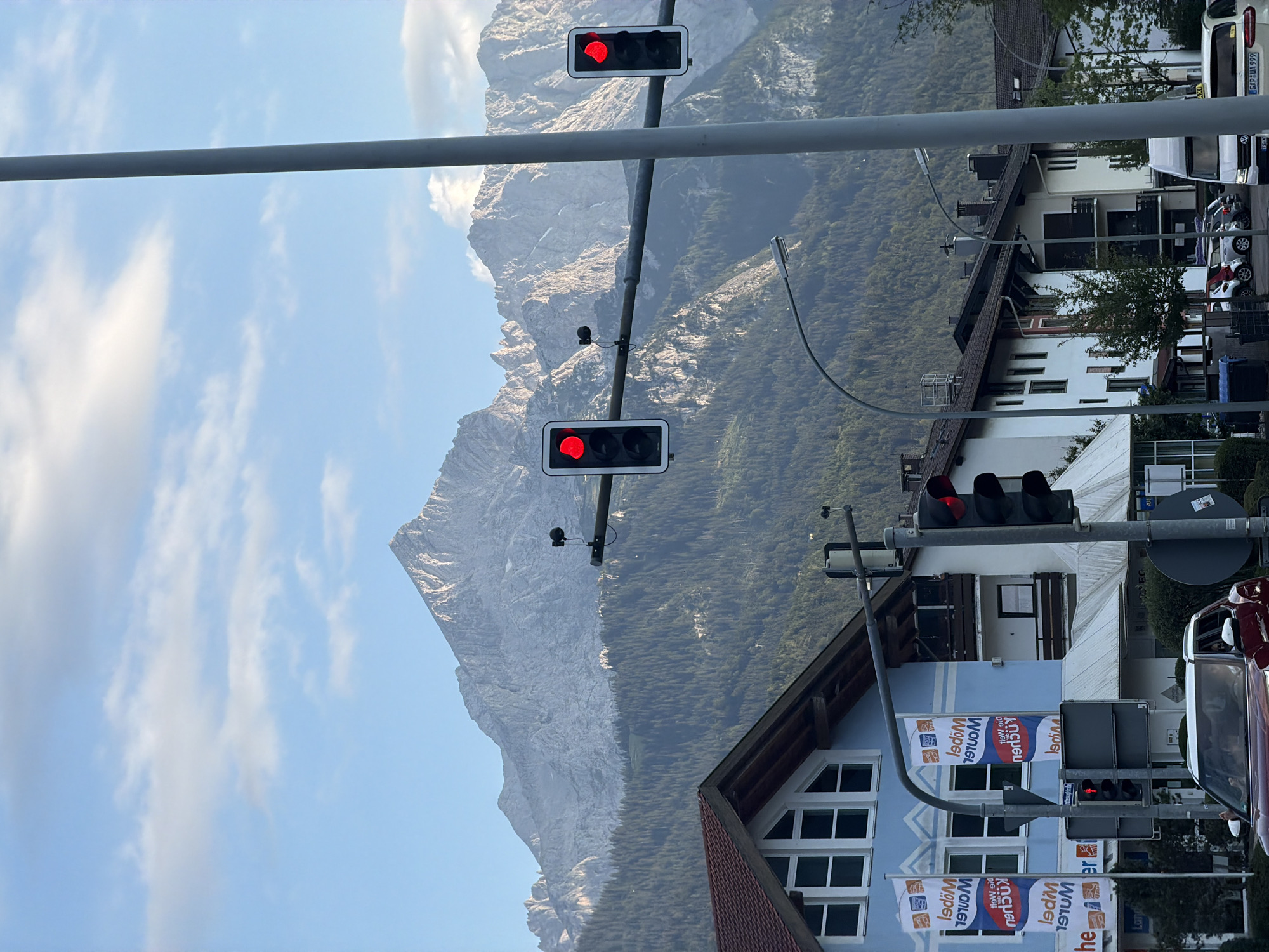 The image captures a view of the Zugspitze mountain range in Garmisch-Partenkirchen, Germany, behind a street scene. In the foreground, traffic lights show a red signal. Buildings with blue and white facades, some with advertising banners, line the street. Several cars are visible, including a red SUV in the lower left. A white van is parked on the right side of the street. The sky is partly cloudy, with the sun illuminating the mountain peaks. The scene appears to be set in the late afternoon or early evening.