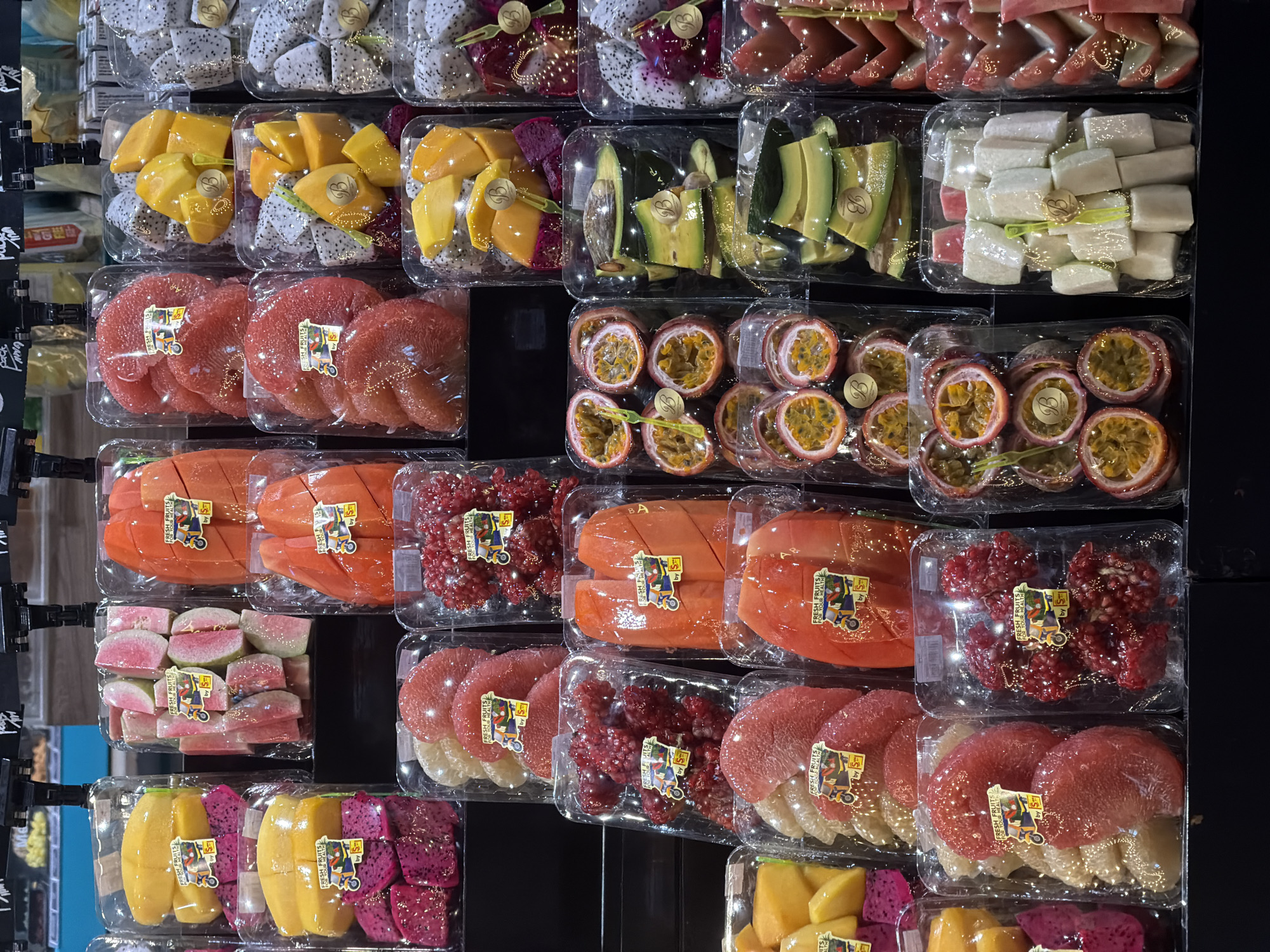 The image displays a retail setting, likely a supermarket or fruit stall, in the city of Ko Samui District, Thailand. The primary focus is on a diverse selection of pre-packaged fresh fruits arranged in clear plastic containers. These containers are stacked on dark shelves, suggesting a tiered display.
The fruits visible include:
* **Dragon fruit**: Both red and white flesh varieties are present, some sliced and some whole.
* **Mango**: Sliced mangoes, predominantly yellow and orange in color.
* **Guava**: Pink-fleshed guavas, cut into wedges.
* **Papaya**: Sliced papaya, orange in color.
* **Pomelo**: Segments of pink grapefruit-like fruit.
* **Pomegranate**: Whole pomegranates and some appearing to be de-seeded.
* **Passion fruit**: Halved passion fruits, revealing their pulp and seeds.
* **Avocado**: Sliced avocado pieces.
* **Rambutan or Lychee**: Sliced fruits with a pinkish-red outer appearance.
* **White, cubed fruit**: Possibly a type of melon or sweet preparation, cut into small rectangular pieces.
Many of the fruit packages feature small, colorful stickers with branding elements, including text like "FRESH FRUITS FOR YOUR HEALTH" and images of a scooter. The lighting suggests an indoor environment, possibly with overhead fluorescent or LED lights typical of retail spaces. No people are visible in the image. The overall impression is one of abundance and fresh produce availability in a commercial setting in Ko Samui.