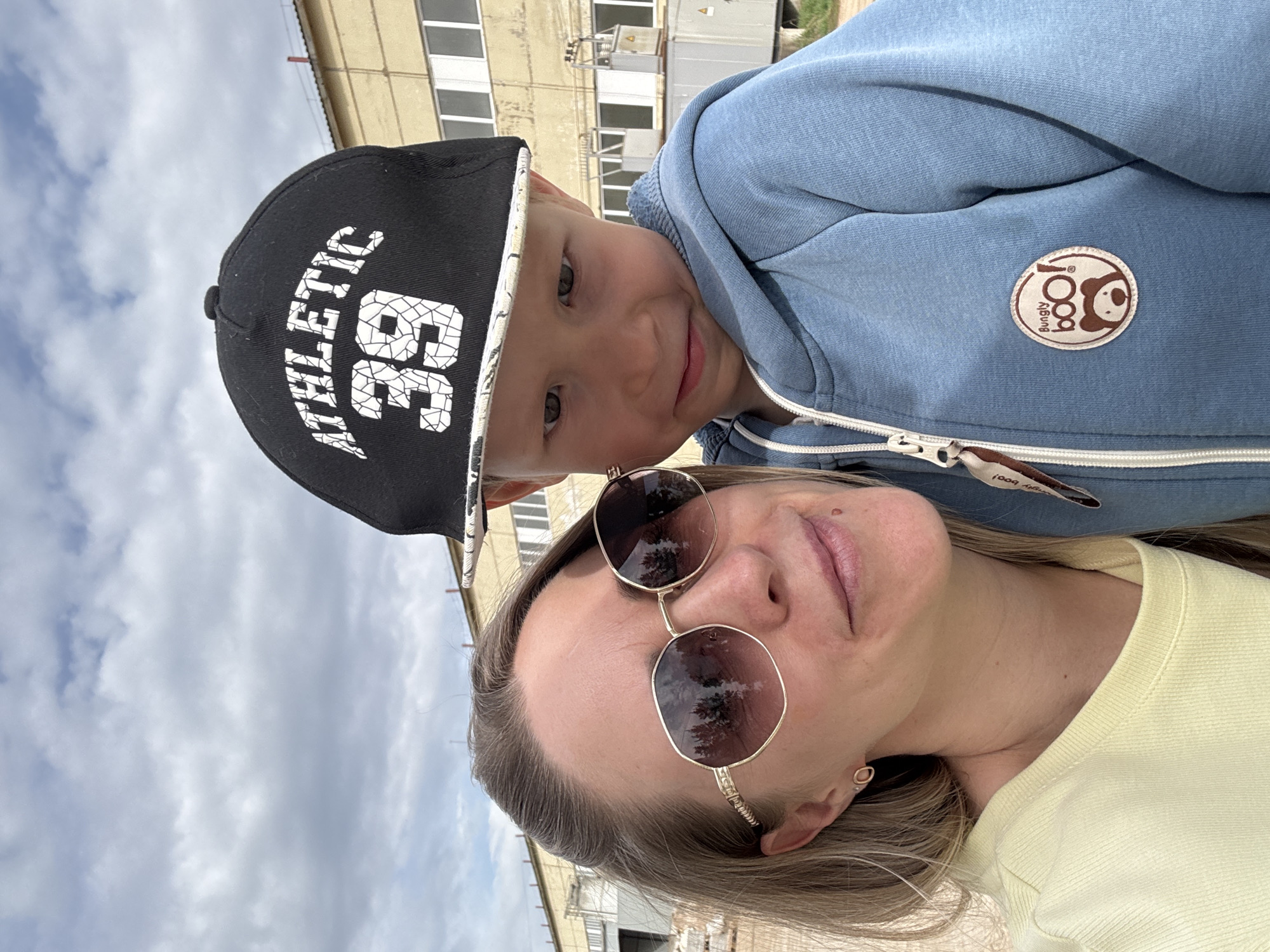 The image shows a selfie of a woman and a young boy. The woman is wearing sunglasses and a yellow shirt. The boy is wearing a blue hoodie and a black baseball cap with white lettering that reads "ATHLETIC 36". The background appears to be an urban environment, with buildings and cloudy skies visible. Based on the building architecture, the photo may have been taken in the city of Chaikovsky, Russia.