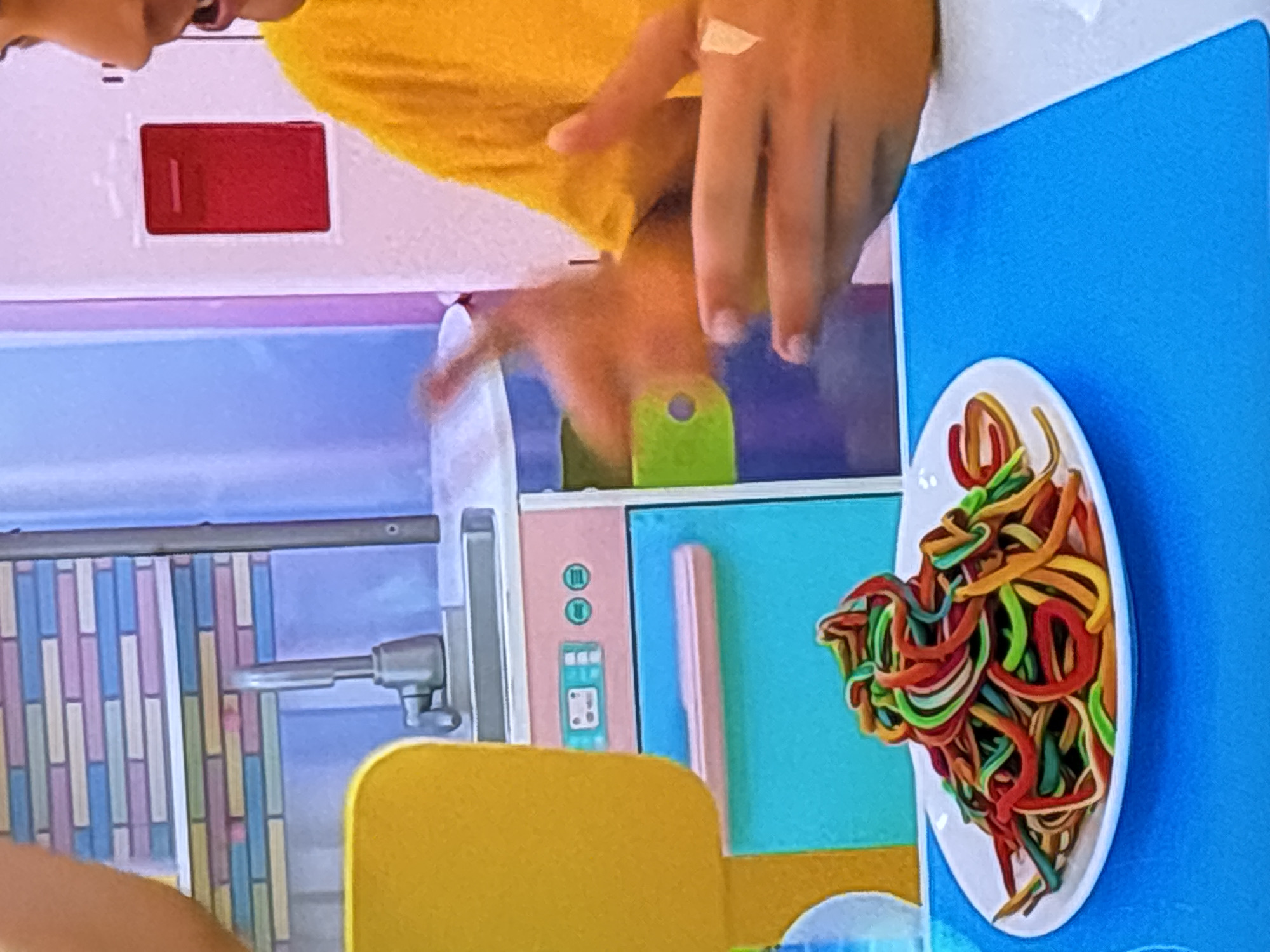 The image captures a close-up view of a child's hands interacting with a pile of colorful, Play-Doh-like spaghetti on a white plate, which sits on a blue placemat. In the background, a toy kitchen playset is visible, featuring a pastel-colored backsplash, a silver faucet, and pink and teal cabinets with a simulated stovetop control panel. To the right, another person, wearing a yellow shirt, is also partially visible, with their hands gesturing. The scene appears to be indoors, possibly in a home or a play area. There is no information in the image to suggest a location in Dubai, United Arab Emirates.