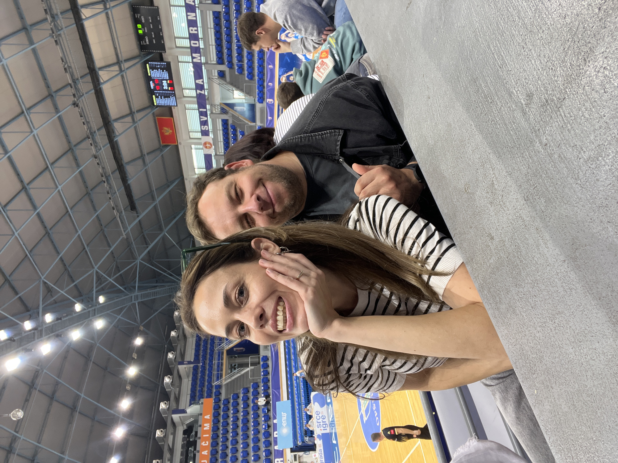 A medium shot captures a smiling woman in a striped shirt with a man behind her giving a thumbs-up, likely at the Topolica Sports Hall in Bar, Montenegro. The backdrop reveals a basketball court with players and blue stadium seating. A scoreboard and the Montenegro flag are visible in the upper part of the frame.