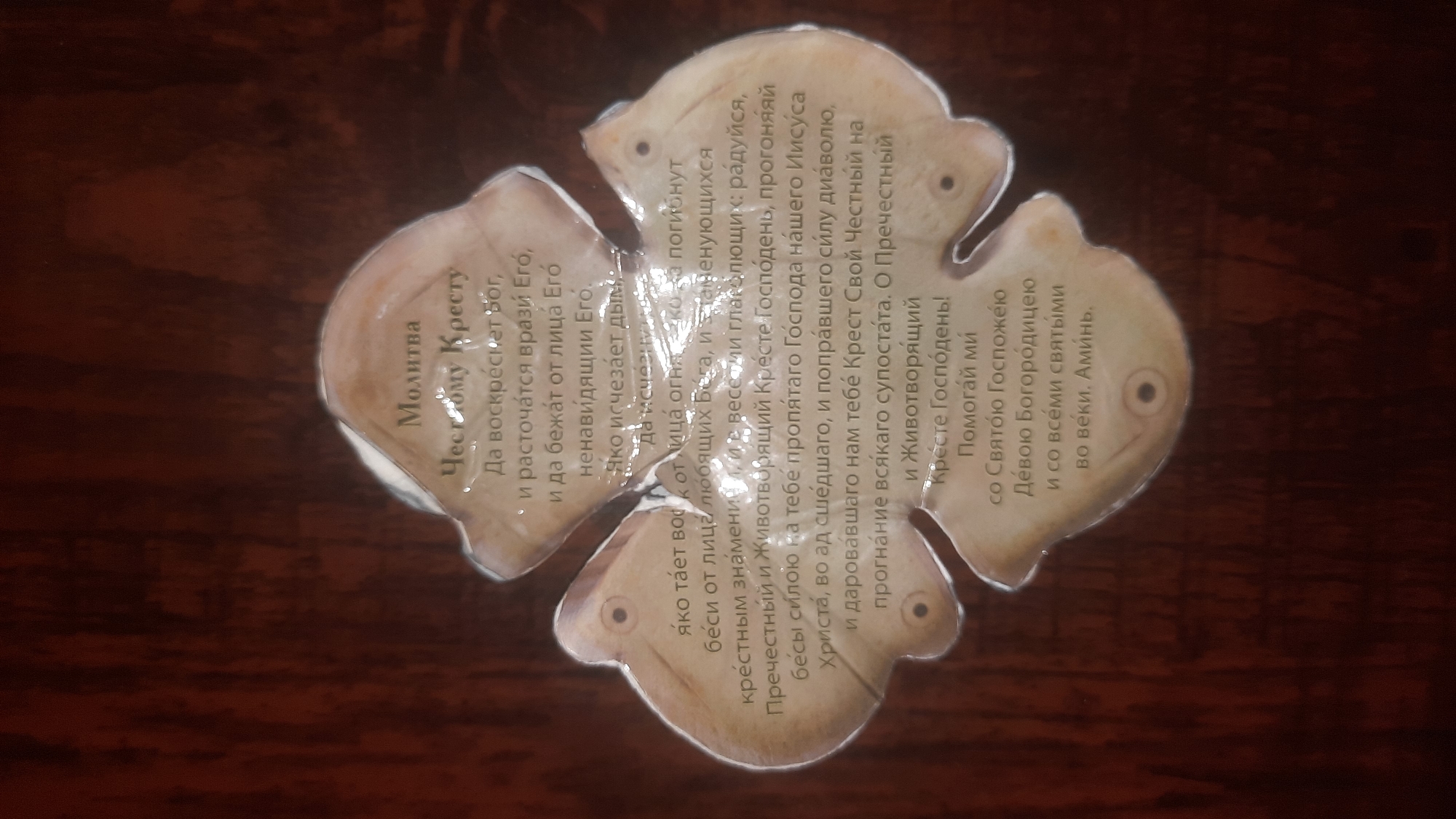 The image shows a cross-shaped object with text on it, placed on a wooden surface. The text is in Russian and appears to be a prayer. The cross-shaped object is made of a light-colored material, possibly paper or thin wood, and has four rounded lobes. The wooden surface is dark brown and has a visible wood grain pattern. There are no people or animals in the image. The location in the city of Shklov, Belarus, is not discernible from the image.