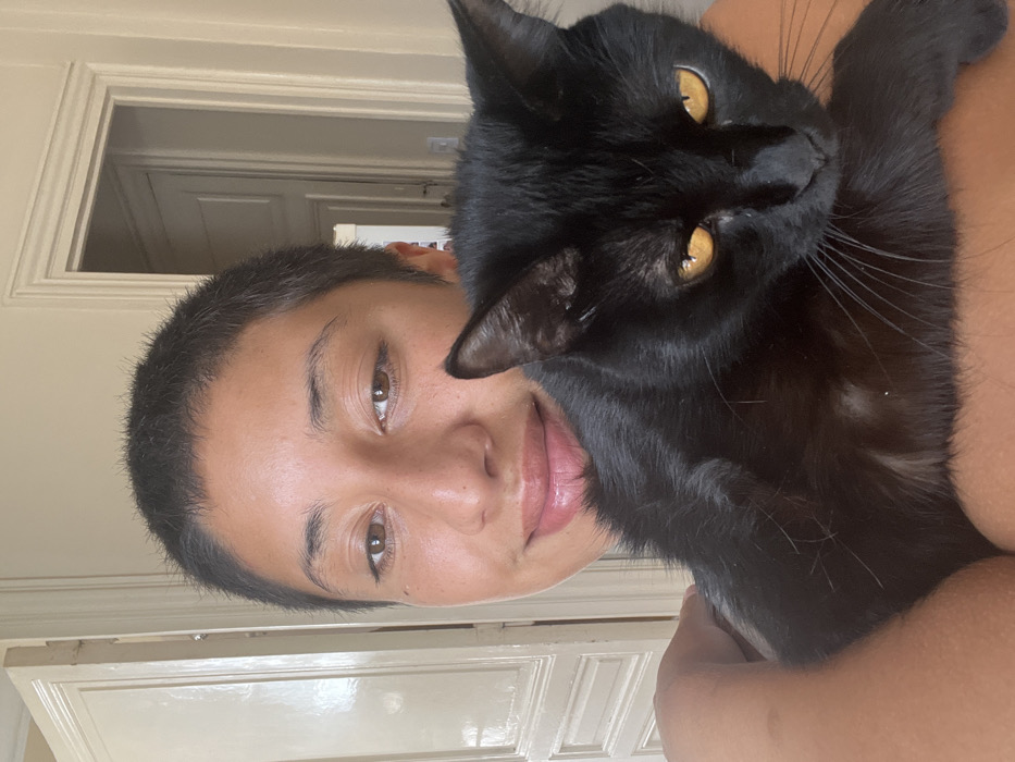 The image depicts a close-up indoor portrait of an individual holding a black cat.
**Individual:** A young adult, likely female, with a short buzz cut hairstyle and medium complexion, is smiling slightly and looking directly at the camera. The individual's bare left shoulder and upper arm are visible, supporting the cat.
**Animal:** A black domestic cat with prominent amber-yellow irises is held against the individual's chest/shoulder. A small patch of white fur is visible on the cat's lower chest/belly. The cat is looking slightly downward and to the right of the frame.
**Interaction:** The individual is holding the cat affectionately in a typical pet-owner embrace.
**Environment/Location:** The background reveals an interior room with light-colored, possibly off-white, walls. Two tall, paneled wooden doors featuring classic decorative molding are present. The door on the left is partially ajar, revealing a darker space beyond. The door frame on the right is also partially visible. This architectural style, characterized by high ceilings (implied by the tall doors) and ornate moldings, is consistent with traditional apartment buildings found in established neighborhoods like Recoleta, Palermo, or Belgrano in Buenos Aires, Argentina. An exact street or building location within Buenos Aires cannot be determined from the visual information provided.
**Lighting:** The scene is illuminated by soft, natural light entering from an unseen source, likely a window.