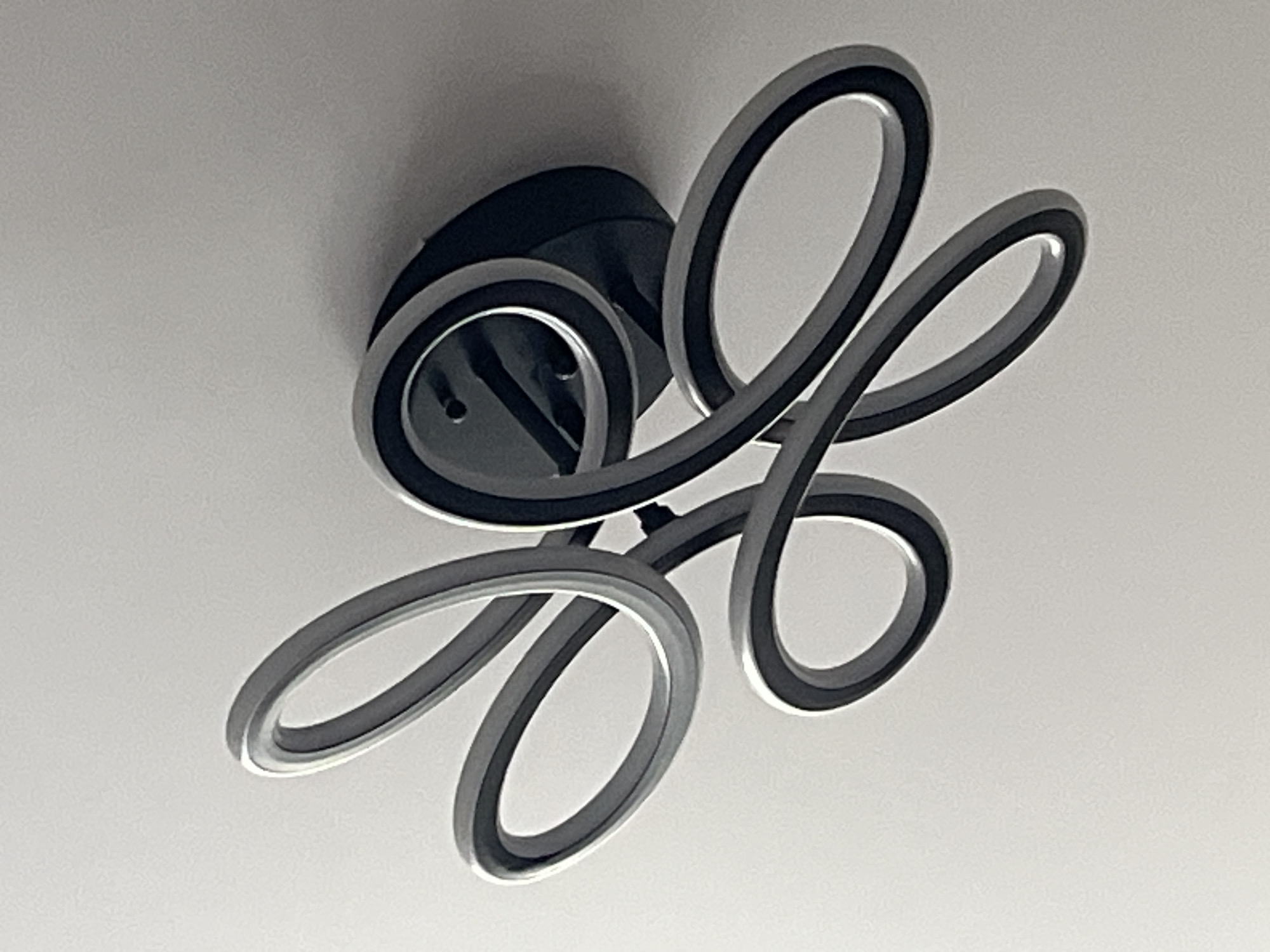 The image shows a modern ceiling light fixture against a plain white ceiling. The fixture consists of a black circular base from which several intertwined, loop-shaped light elements extend. These loops are edged with a dark band and have a lighter grey inner surface. The location is not possible to determine from the image.