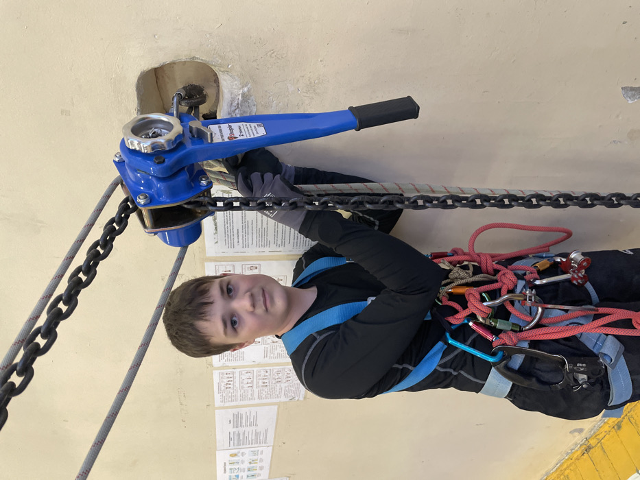 An adolescent male stands indoors in what appears to be a training facility in Sarapul, Russia. He is equipped with a blue and grey full-body harness adorned with multiple carabiners and red and grey ropes, worn over a black long-sleeved shirt and dark pants. His hands are covered by grey and black gloves. The individual is positioned to the left of a blue "Steupler 2 tonne" lever hoist, which is anchored to a light beige wall. A heavy metal chain passes through the hoist and descends vertically; the male's gloved right hand is resting on this chain. A grey rope also descends vertically, parallel to the chain. To the right of the hoist, a rough, rectangular opening is present in the wall. The background features several documents with diagrams and Russian text, including "Порядок работ на высоте" (Order of work at height), taped to the wall. A yellow line is visible on the floor in the lower-left portion of the frame. The male maintains a neutral expression, looking directly forward.