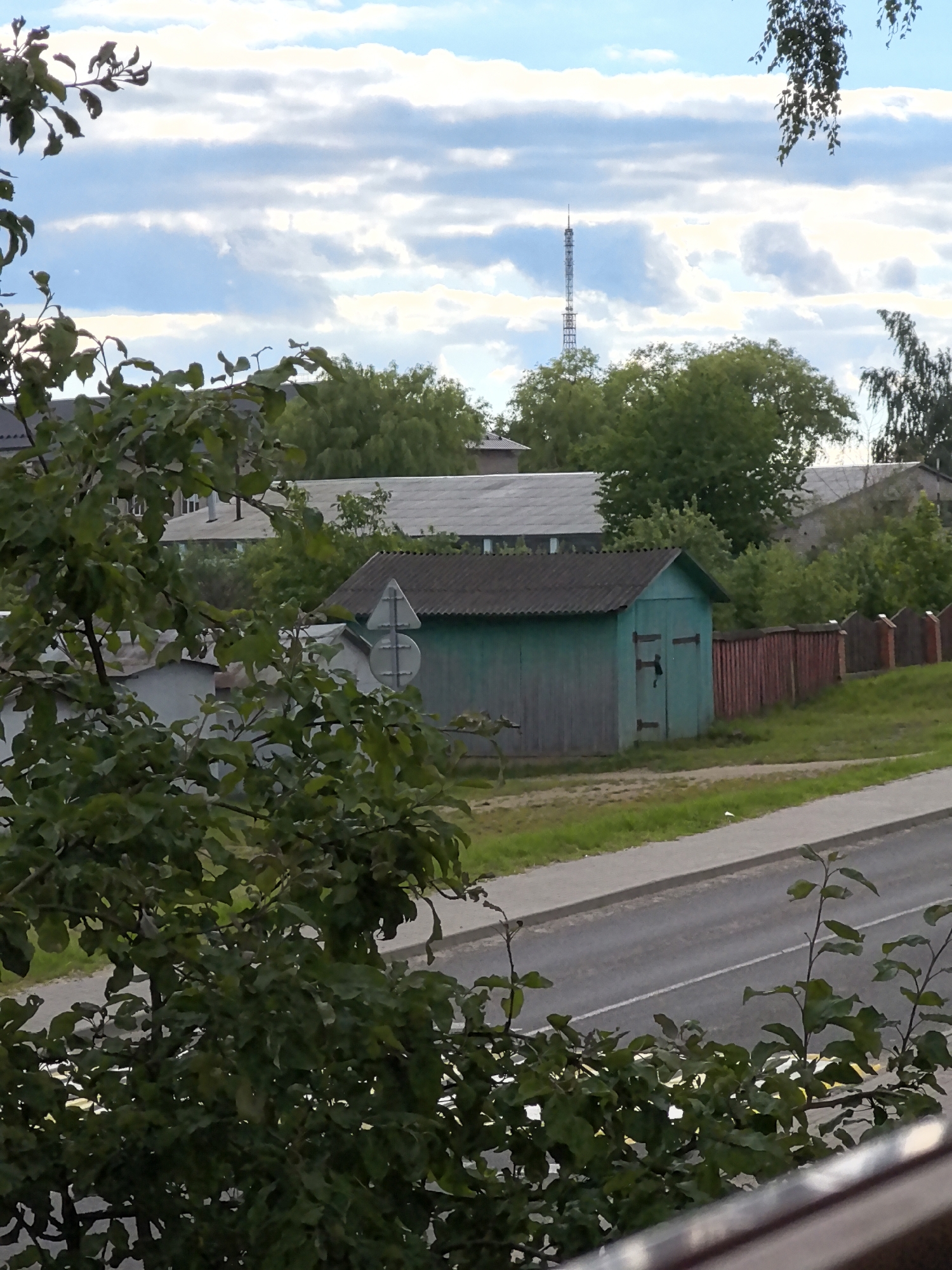 A tall, lattice radio tower is visible in the distance, rising above a cluster of trees and buildings. In the foreground, a vibrant green, wooden shed with a dark roof stands beside a red wooden fence. A road with white and yellow markings runs in front of the shed and fence, bordered by a grassy area. The sky is a mix of blue and white clouds, with a few leafy branches from a tree obscuring parts of the view. The scene appears to be an outdoor setting, possibly in a rural or suburban area, with a clear, bright sky overhead.