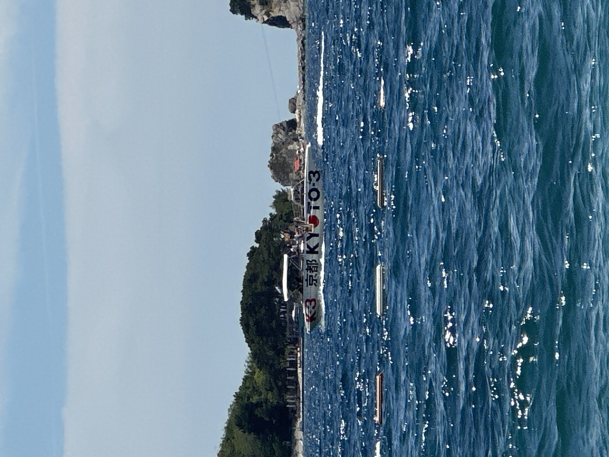 This image depicts a sunny day at sea, likely off the coast of Budva, Montenegro. In the mid-ground, a white speedboat with the markings "K3 京都 KYOTO-3" is moving from left to right, leaving a white wake in the deep blue, choppy water. The boat appears to be carrying passengers. In the background, a lush, green, tree-covered hillside with some structures is visible on the left, transitioning to rocky outcrops and cliffs on the right. The sky is a bright, clear blue with some wispy white clouds. In the foreground, several long, rectangular objects, possibly buoys or floating platforms, are partially submerged in the water, marked with alternating dark and light stripes.