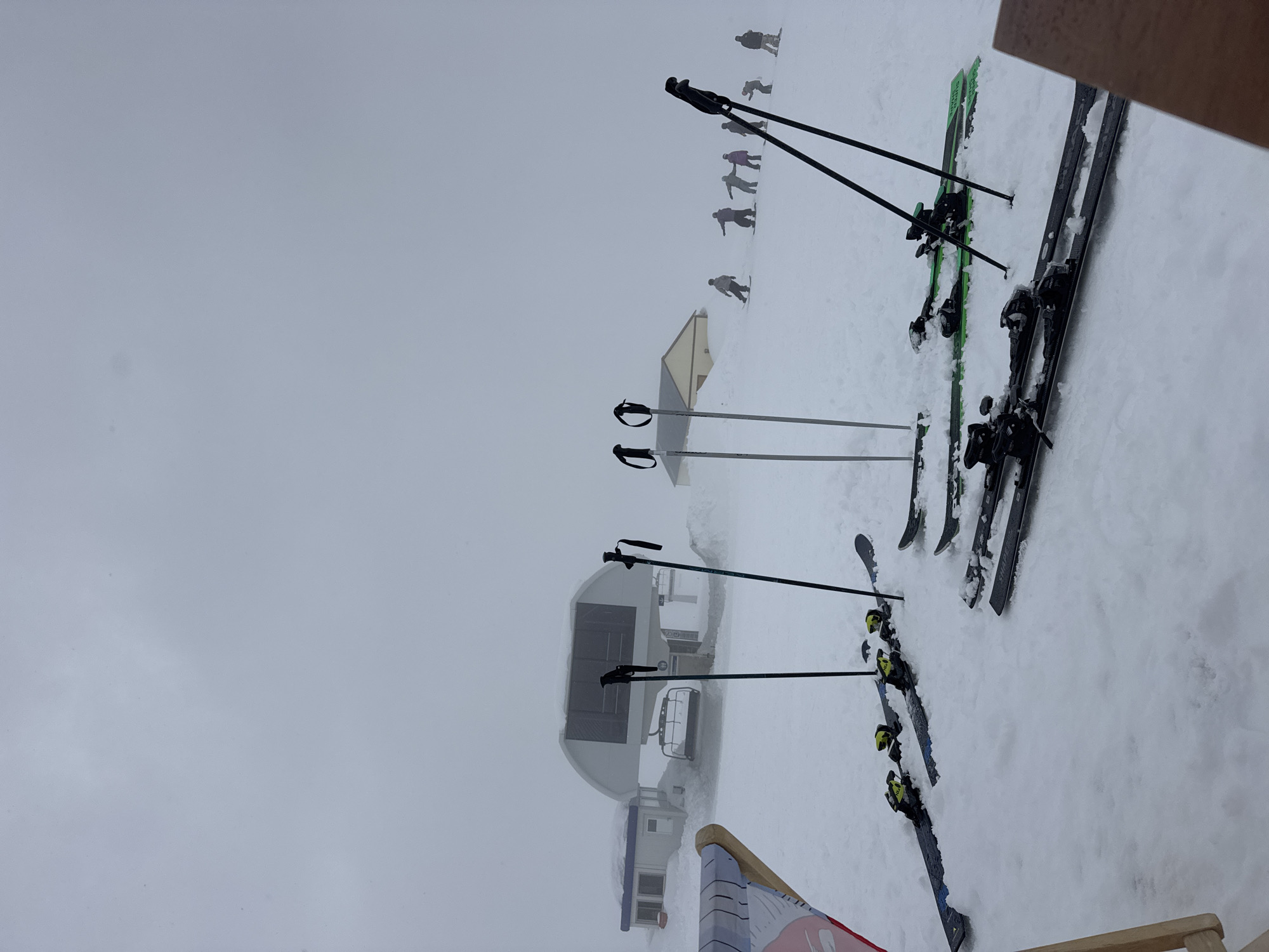 The image shows a snowy scene at what appears to be a ski resort. Three pairs of skis and poles are standing upright in the snow. In the background, there is a ski lift station and several people skiing or snowboarding in the distance on a slope. The overall environment suggests a cold, overcast day due to the fog.