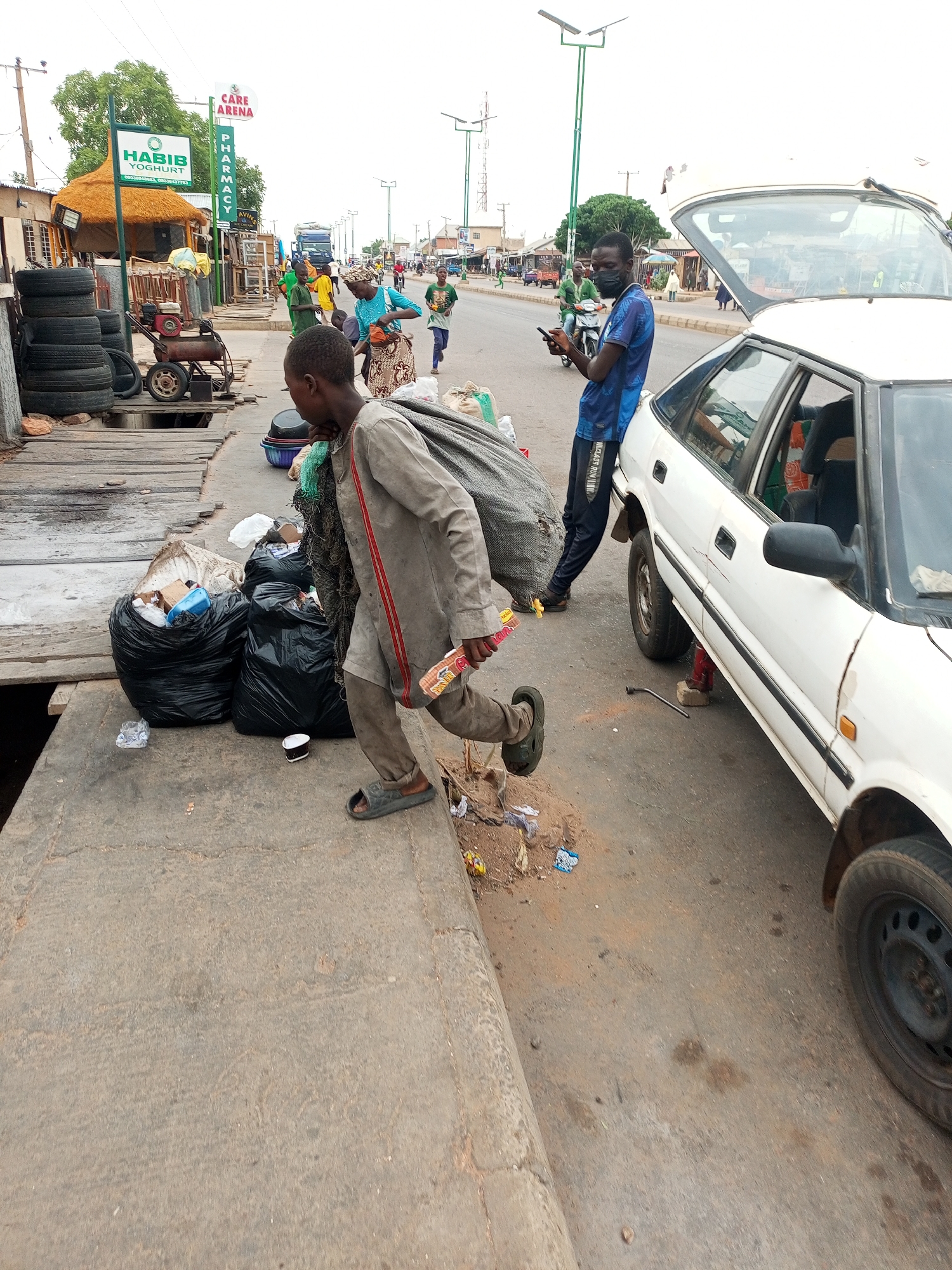 The image shows a street scene in Gombe, Nigeria. A young man wearing a grey long-sleeved shirt with a red stripe, brown pants, and sandals is carrying a large, grey sack. He is holding a wrapped package in his right hand and is walking from a platform towards the street. Next to the platform are several black trash bags filled with garbage.
Beside the young man, a white car is jacked up, with the front passenger side wheel removed. A man wearing a blue shirt, black pants, and a black face mask is standing next to the car, looking at his phone. The car's trunk is open.
In the background, there is street traffic consisting of pedestrians and vehicles. On the left side of the image, there is a business with a sign that says "HABIB YOGHURT" and "CARE ARENA PHARMACY". There are tires and a small gas-powered engine on the platform near the trash bags. The sky is overcast.