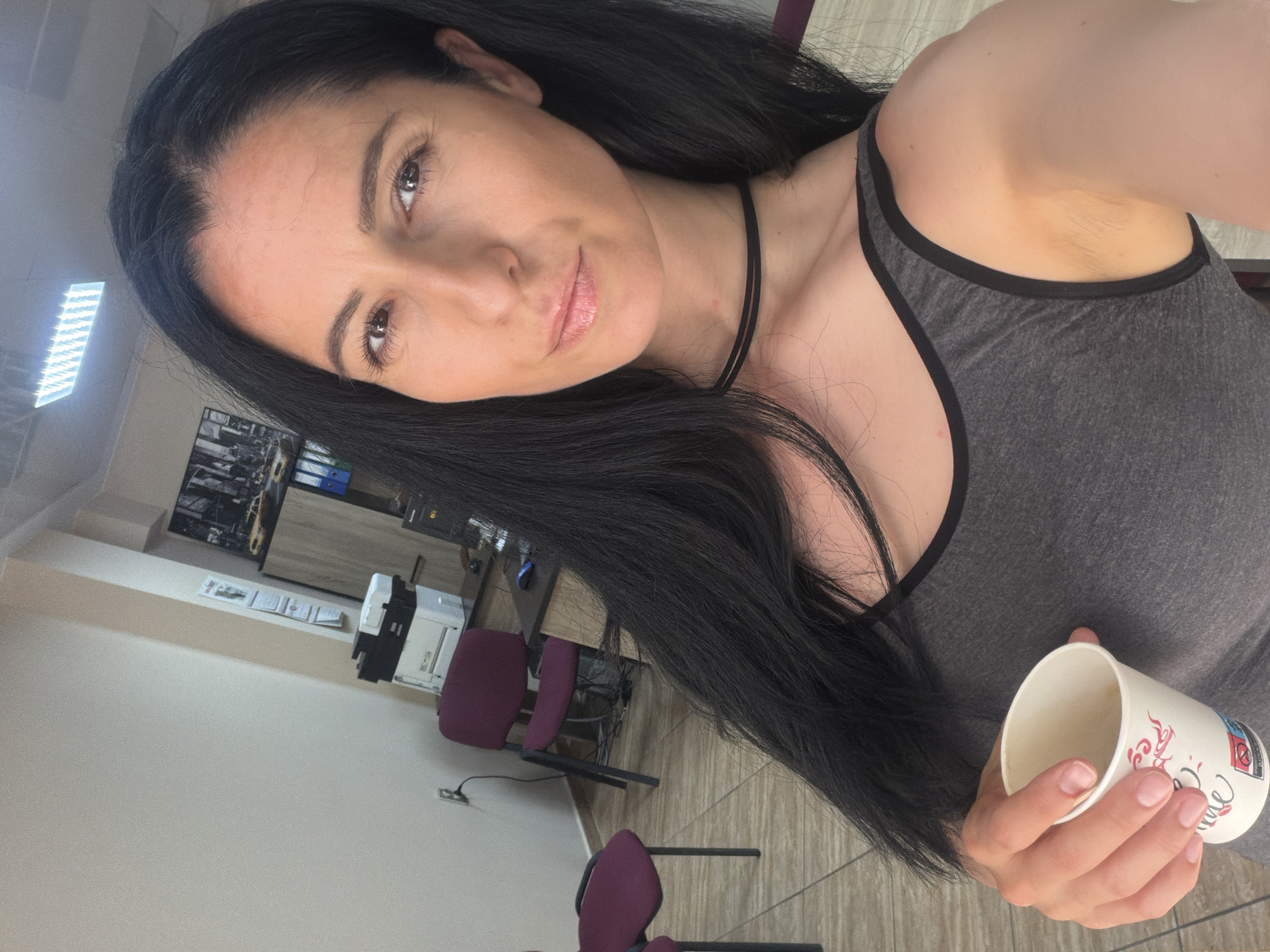 A woman with long, dark hair is taking a selfie in an office environment. She is wearing a grey tank top and a black choker necklace. She is holding a white paper cup in her left hand. The background shows office furniture, including chairs, a desk with a printer and computer equipment, and filing cabinets. The lighting suggests an indoor setting, possibly during the day. The image appears to be taken in Pleven, Bulgaria, based on the location tag.