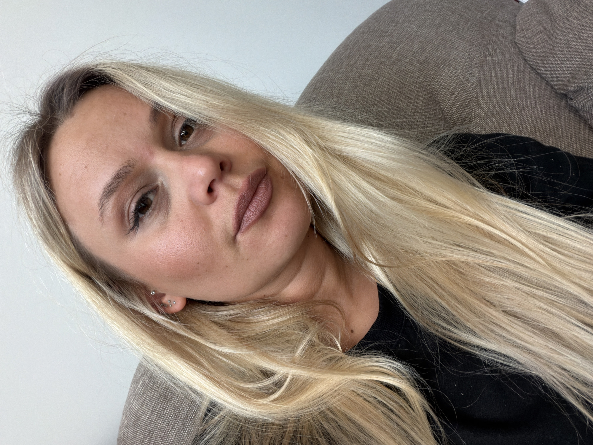 A woman with blonde hair is reclining on a grey couch, wearing a black top. Her head is tilted, and she is looking at the camera. She has multiple piercings in her left ear. The background consists of a white wall. There is no indication of the location being Sarapul, Russia.