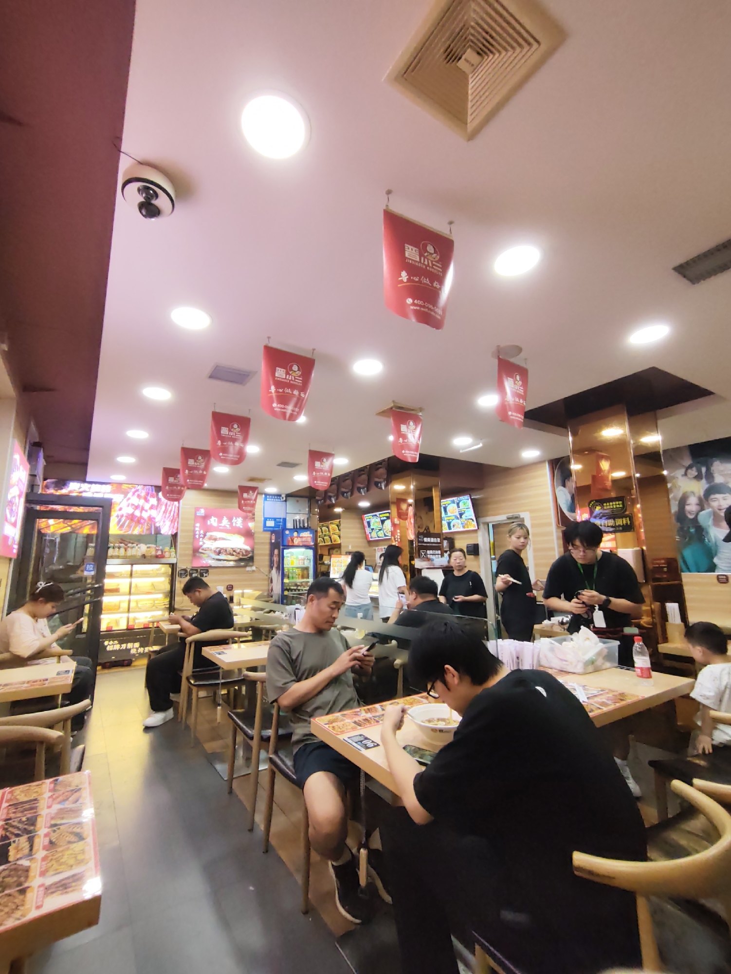The image depicts the interior of a brightly lit restaurant in Aihui, China. Red banners with Chinese characters hang from the ceiling. Several patrons are seated at tables, many of them engaged with their mobile phones. Some are eating, with bowls and food visible on the tables. In the background, there are display cases with food items and a refrigerator stocked with beverages. A security camera is mounted on the ceiling. The overall atmosphere suggests a casual dining establishment.