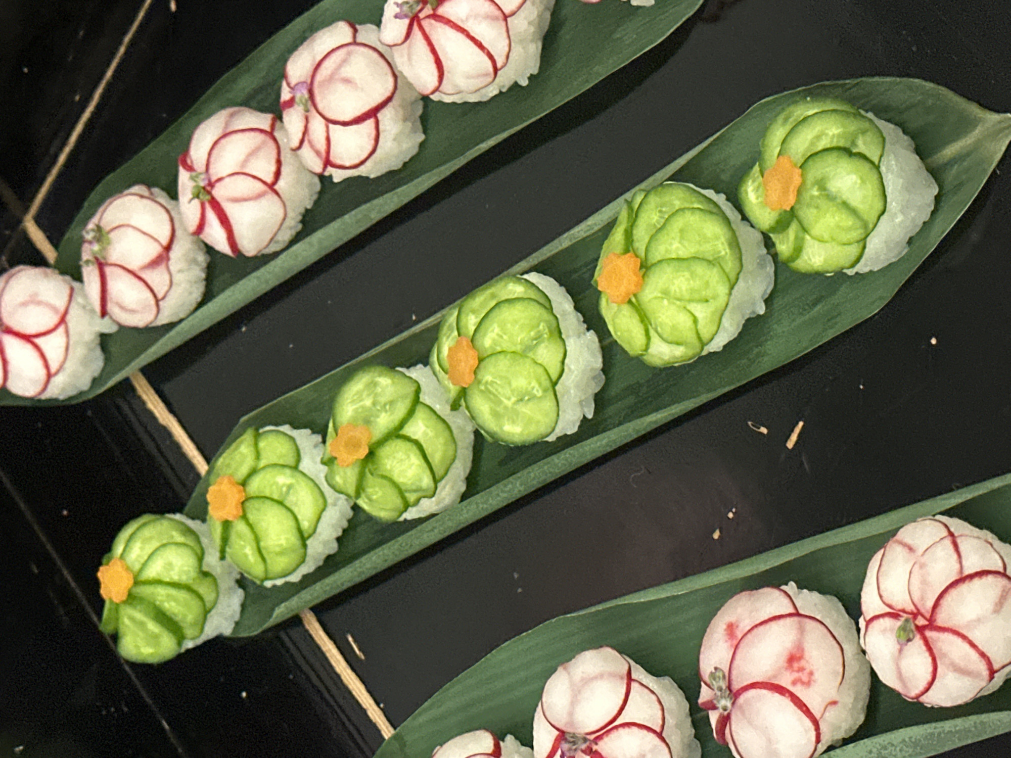A close-up, high-angle view displays multiple sushi-like food items arranged in rows on green leaves over a dark, reflective surface. Three parallel rows of these items are visible. The two outer rows, on the left and right, consist of white rice mounds topped with thinly sliced white radish petals, which are edged in red; some of these pieces are centrally adorned with a small purple garnish. The central row features white rice mounds topped with overlapping green cucumber slices, arranged in a petal formation, each centrally garnished with a small orange granular element. The food items rest on elongated, narrow green leaves, resembling bamboo leaves, which are laid upon a uniform black or dark brown reflective surface, possibly a lacquered tray. The specific location within the city of London, United Kingdom, cannot be determined from the visual information provided in the image.